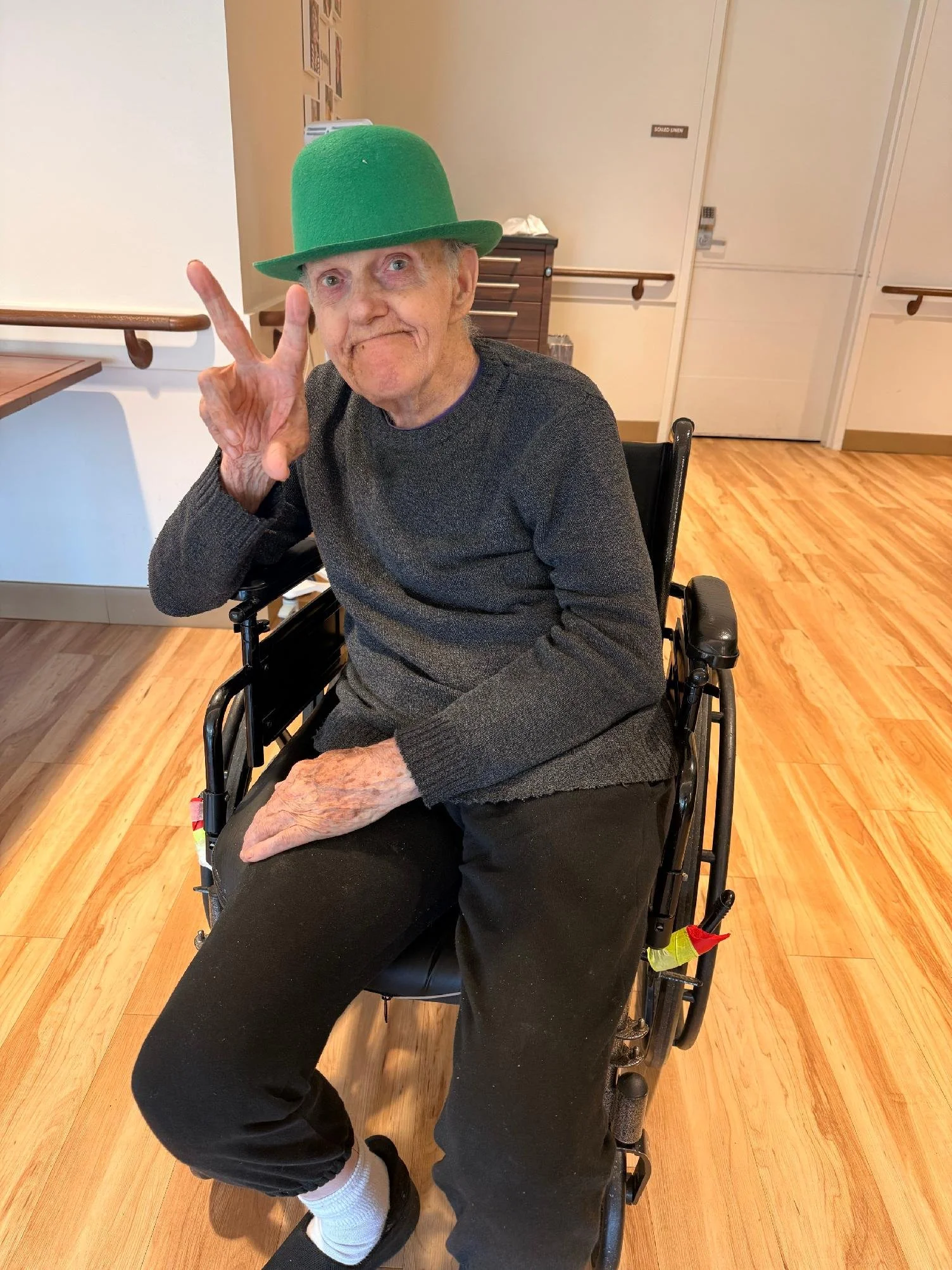An elderly man with gray hair and a mustache sitting in a wheelchair, giving a peace sign with his right hand. He is wearing a green felt hat, a gray sweater, black pants, white socks, and black shoes. The background shows a hospital or care facility