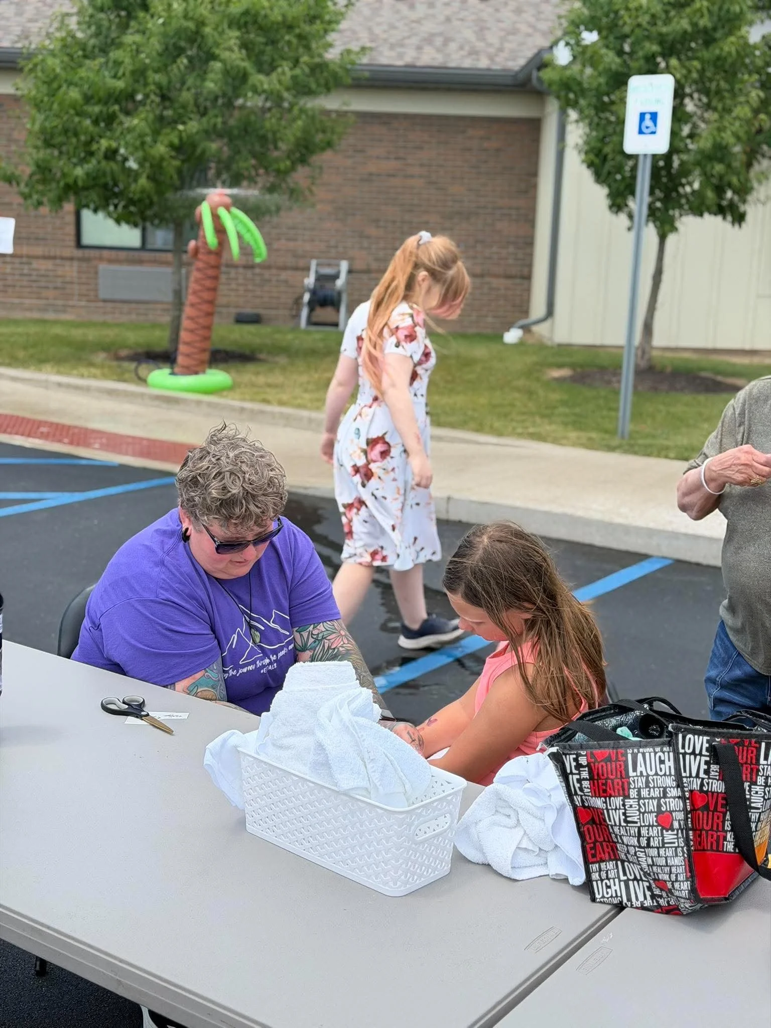 A group of people gathered around a table outdoors, with one person receiving a tattoo on their arm, and supplies like paper towels and scissors on the table. In the background, a brick building, trees, and a parking lot with a handicapped sign are v