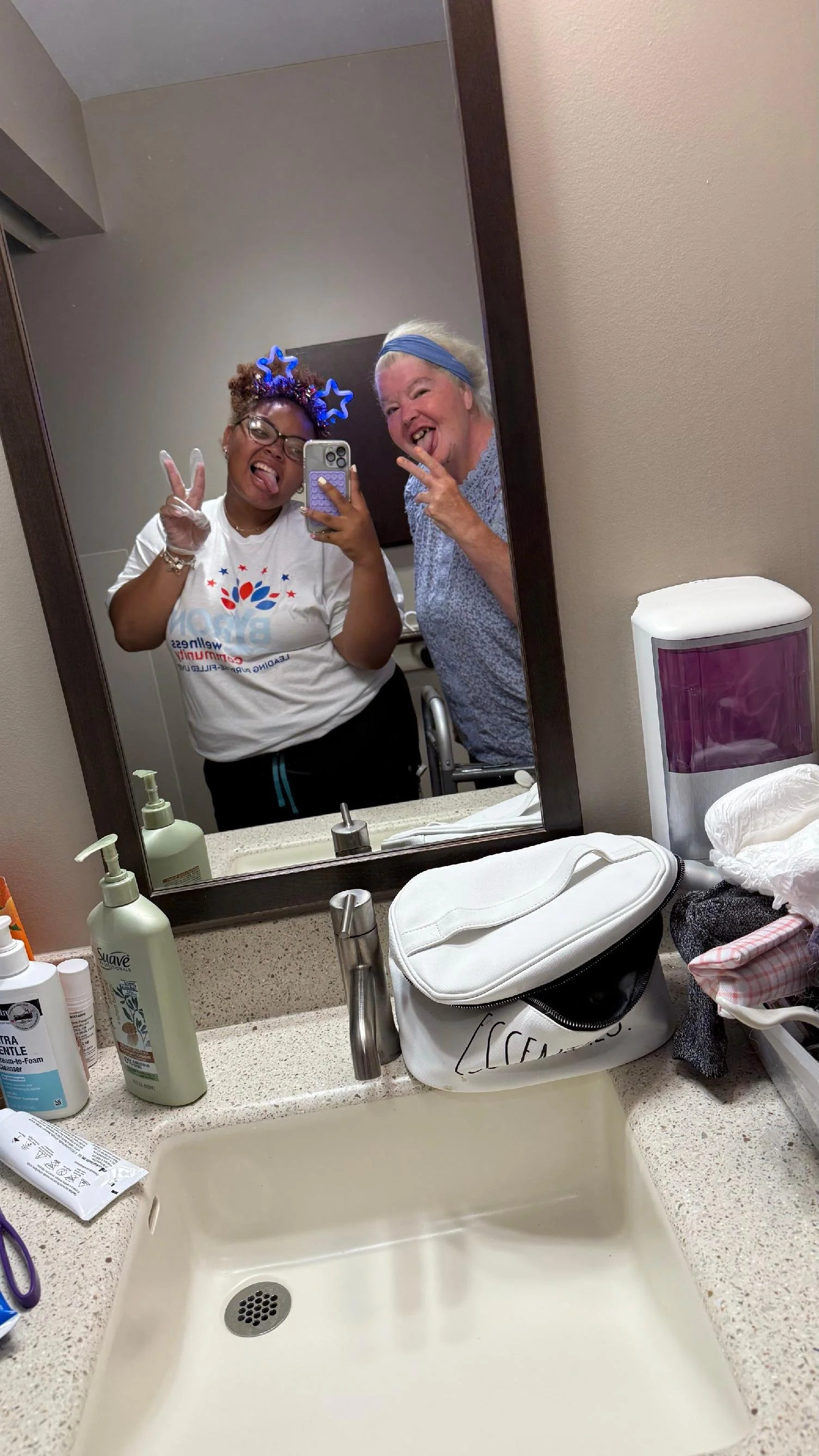 Two women taking a selfie in front of a bathroom mirror, making silly faces and peace signs. One woman has glasses, a glow star headband, and is wearing a white shirt. The other woman has a blue headband and is wearing a light blue top. The bathroom 