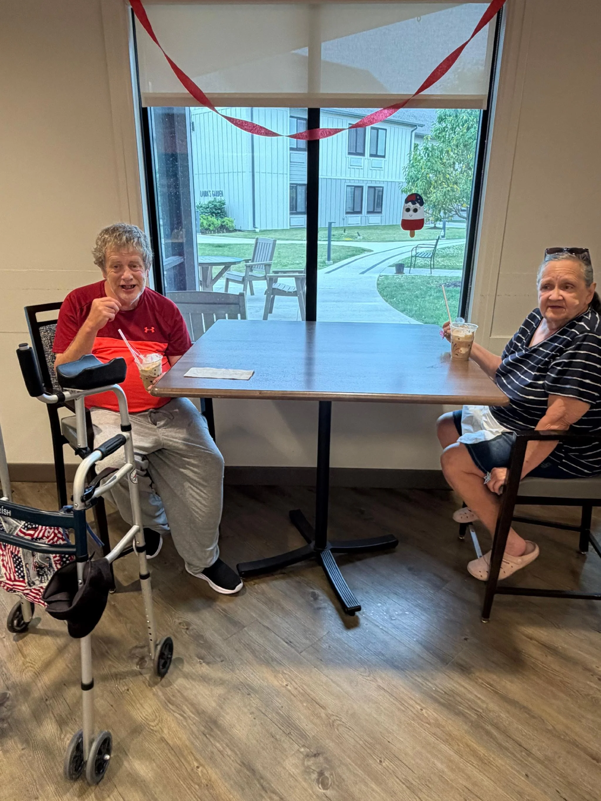 Two elderly women sitting at a table in a room with a window showing an outdoor patio with chairs and a tree. They are holding drinks with straws; one has a walker beside her, and both women are smiling or talking.