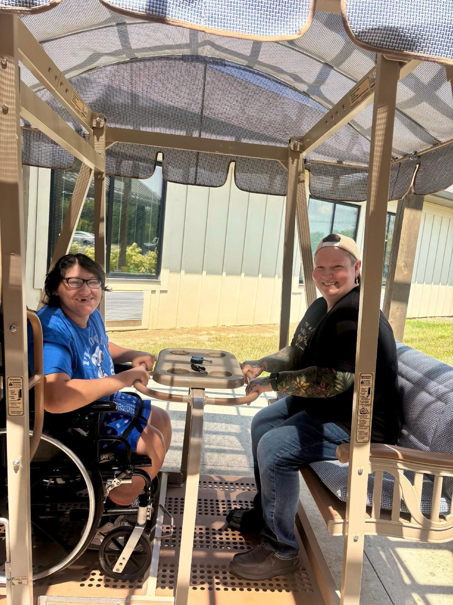 Two people sitting inside a small, open-sided vehicle with a canopy, smiling at the camera. One person is a woman with glasses, sitting in a wheelchair, and the other is a man with a hat, sitting on a bench seat. They are outdoors near a building wit