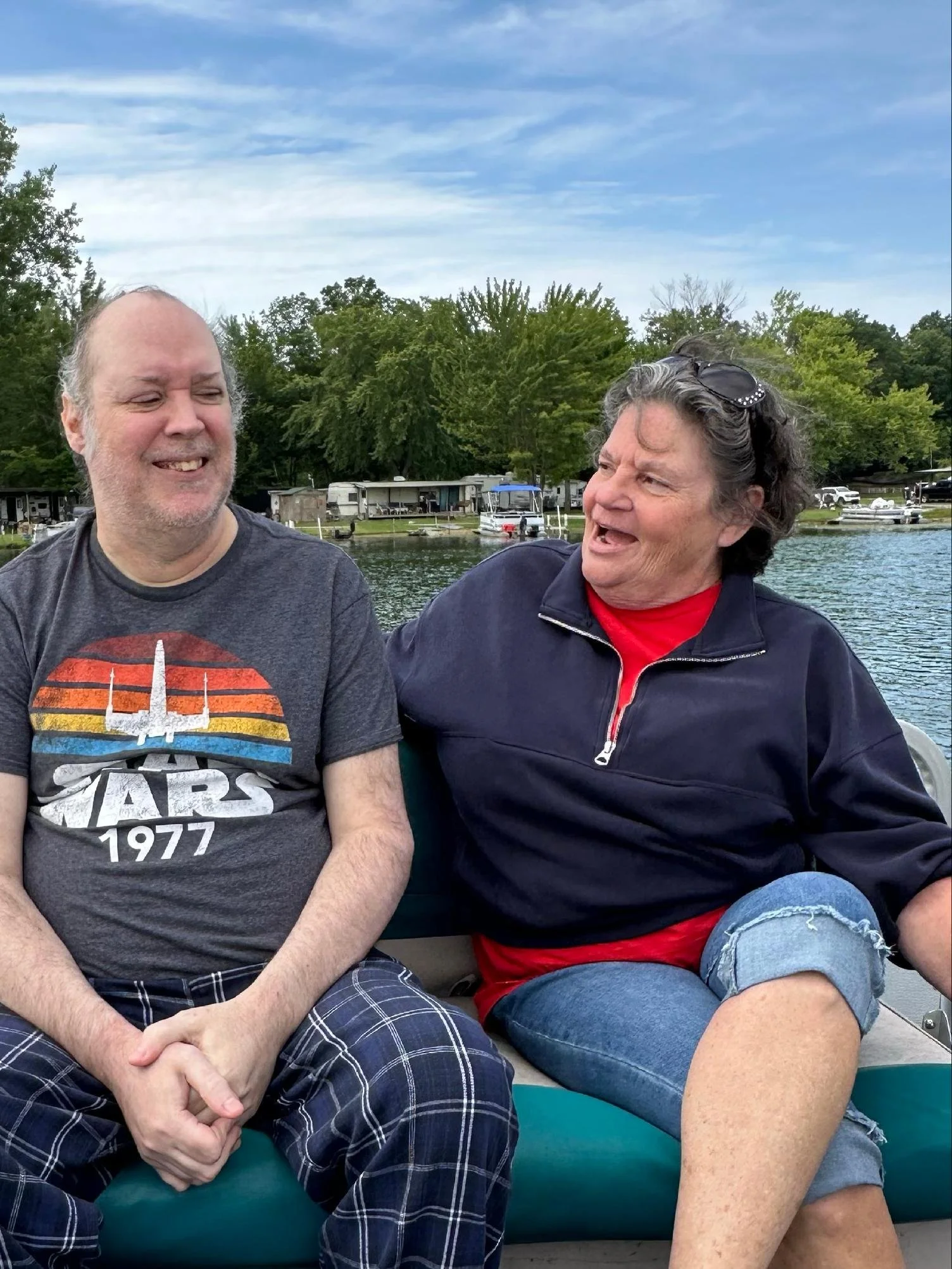 Two people sitting by a lake, smiling and talking, with boats and green trees in the background.