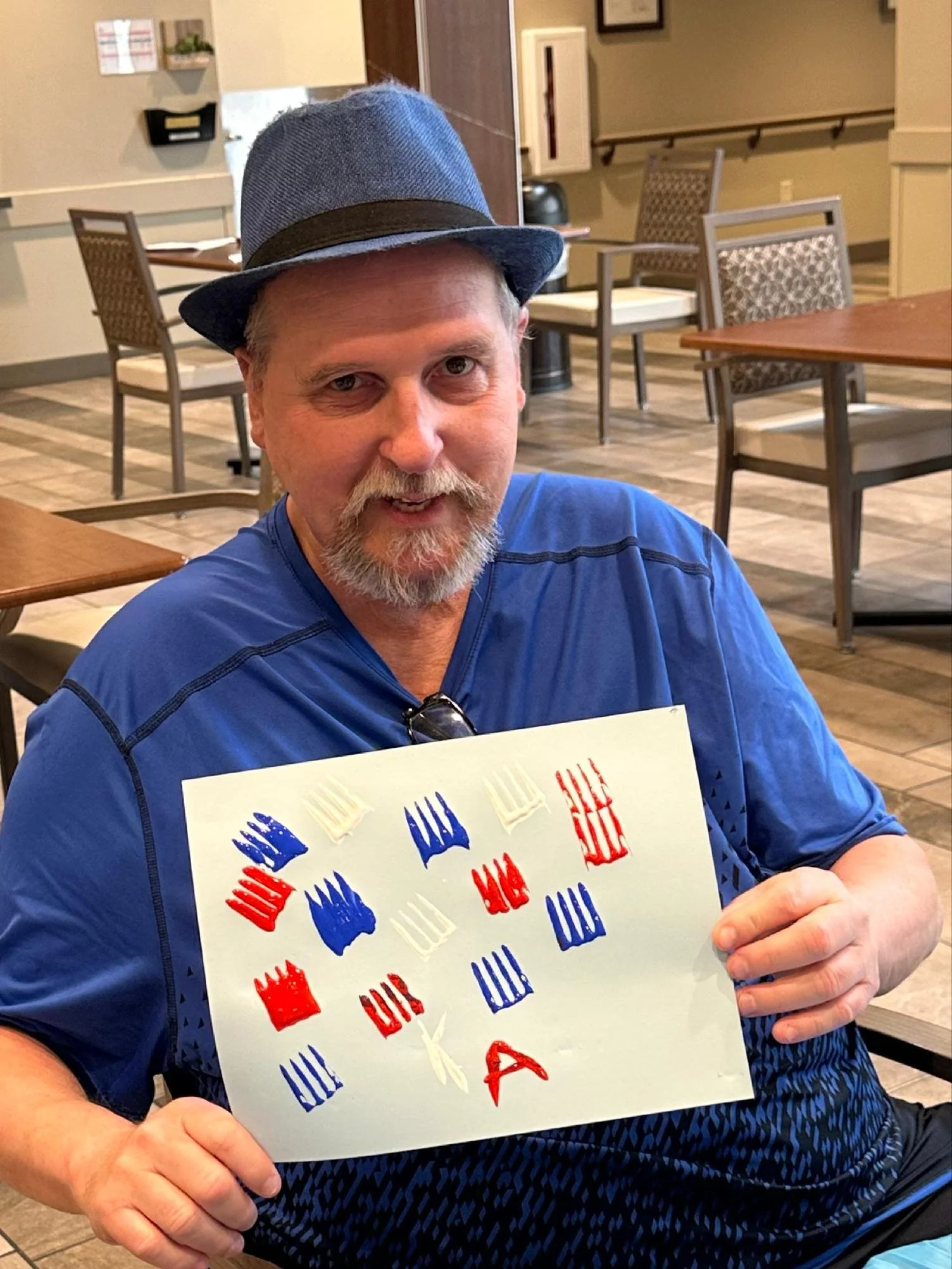 A man with a beard and mustache wearing a blue shirt and a gray hat sitting in a restaurant, holding a piece of paper with red, white, and blue fork and spoon stamps.