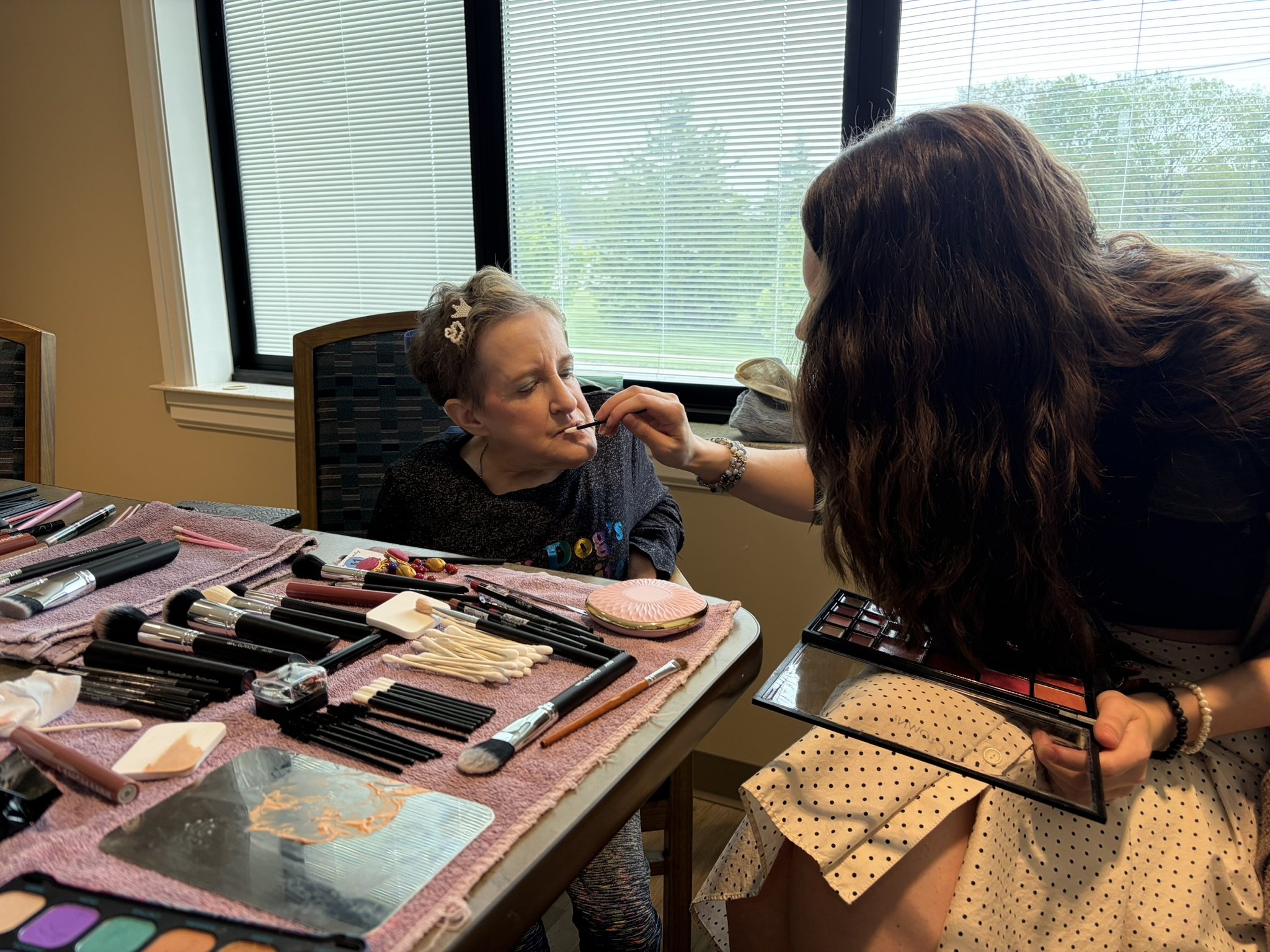 A woman with long brown hair applying makeup to an older woman with short gray hair. The older woman is sitting in a chair while the woman with long hair applies makeup. The table in front has various makeup brushes, tools, and products organized on 