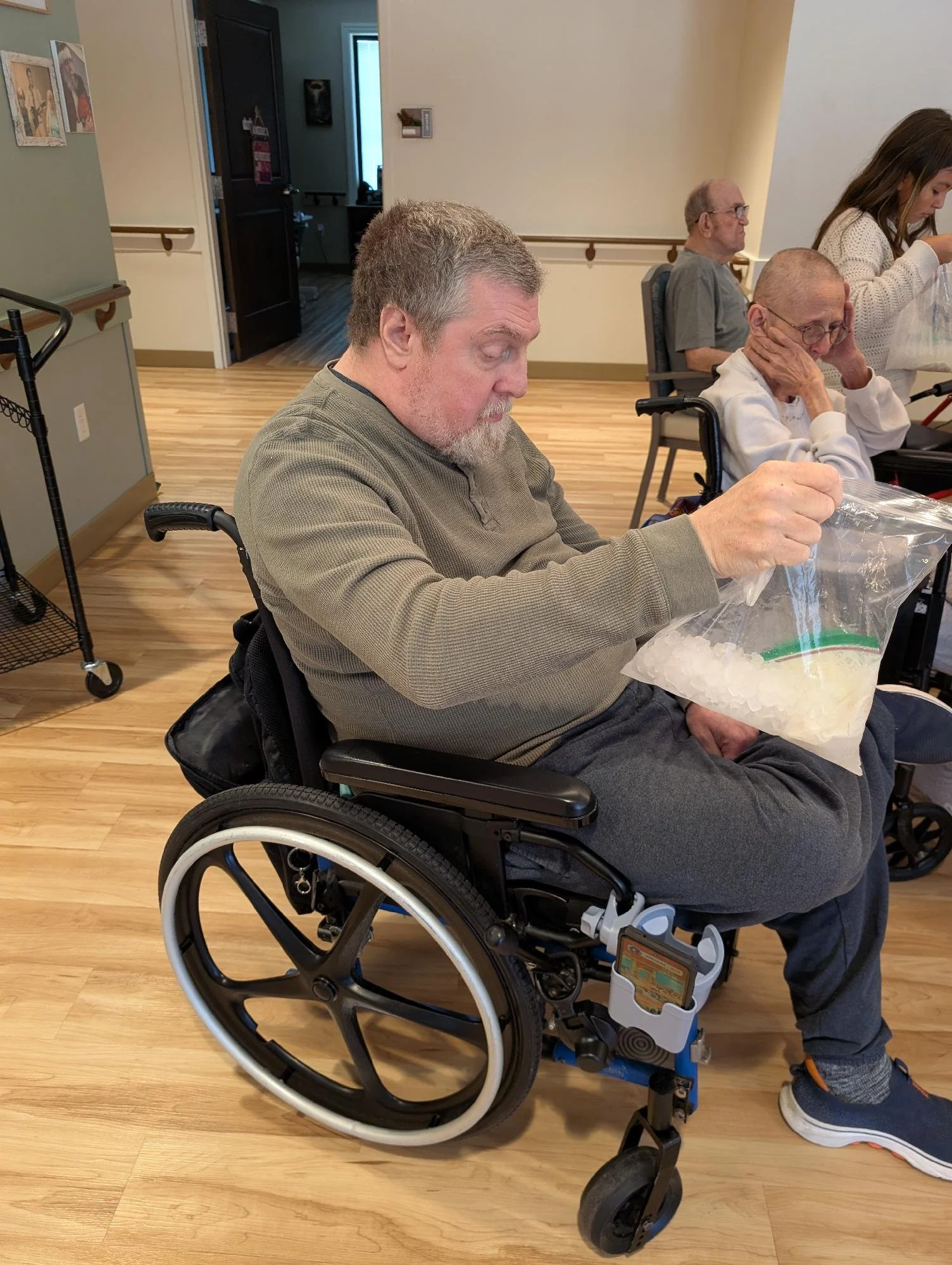 An elderly man in a wheelchair holding a plastic bag with ice, with an older woman sitting next to him holding her head, in a room with wood flooring and beige walls, and a young girl standing nearby.
