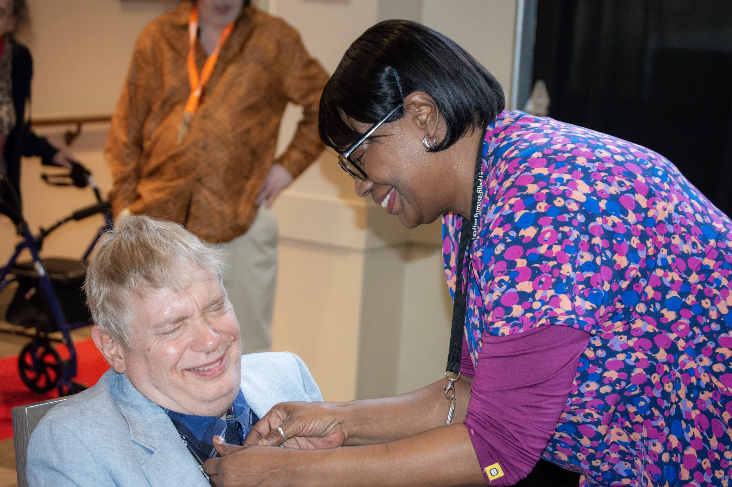 A woman in a colorful dress smiling warmly as she adopts a caring posture towards an elderly man with white hair, who is seated and smiling with closed eyes. Two other people are in the background, observing the interaction.