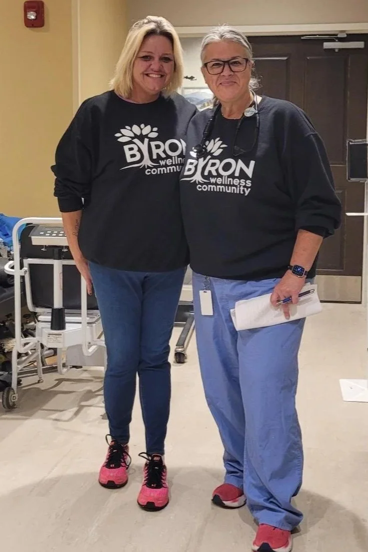 Two women standing together, smiling, wearing matching black sweatshirts with 'BRON wellness community' logos. They appear to be in a healthcare or community center setting.