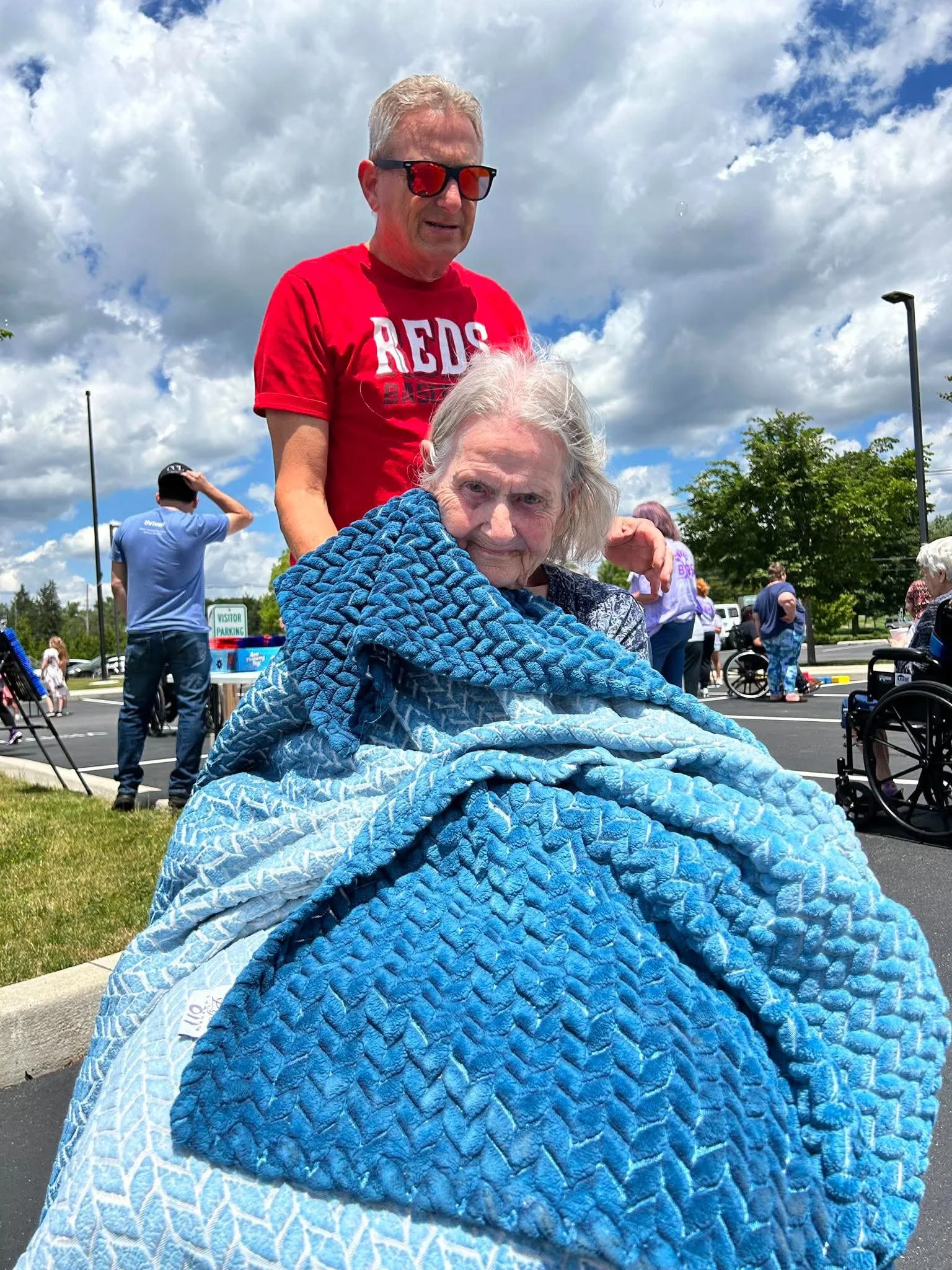 An elderly woman in a wheelchair wrapped in a blue quilt blanket with a textured pattern and sitting outside on a sunny day, with a middle-aged man standing behind her wearing sunglasses and a red shirt with white letterings.
