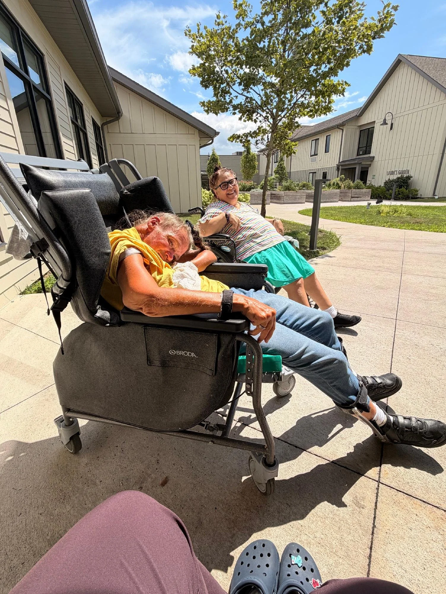 Two elderly women sitting outdoors on a sidewalk, one in a wheelchair smiling and the other smiling and leaning back. The scene is sunny with a blue sky and green trees and plants nearby.