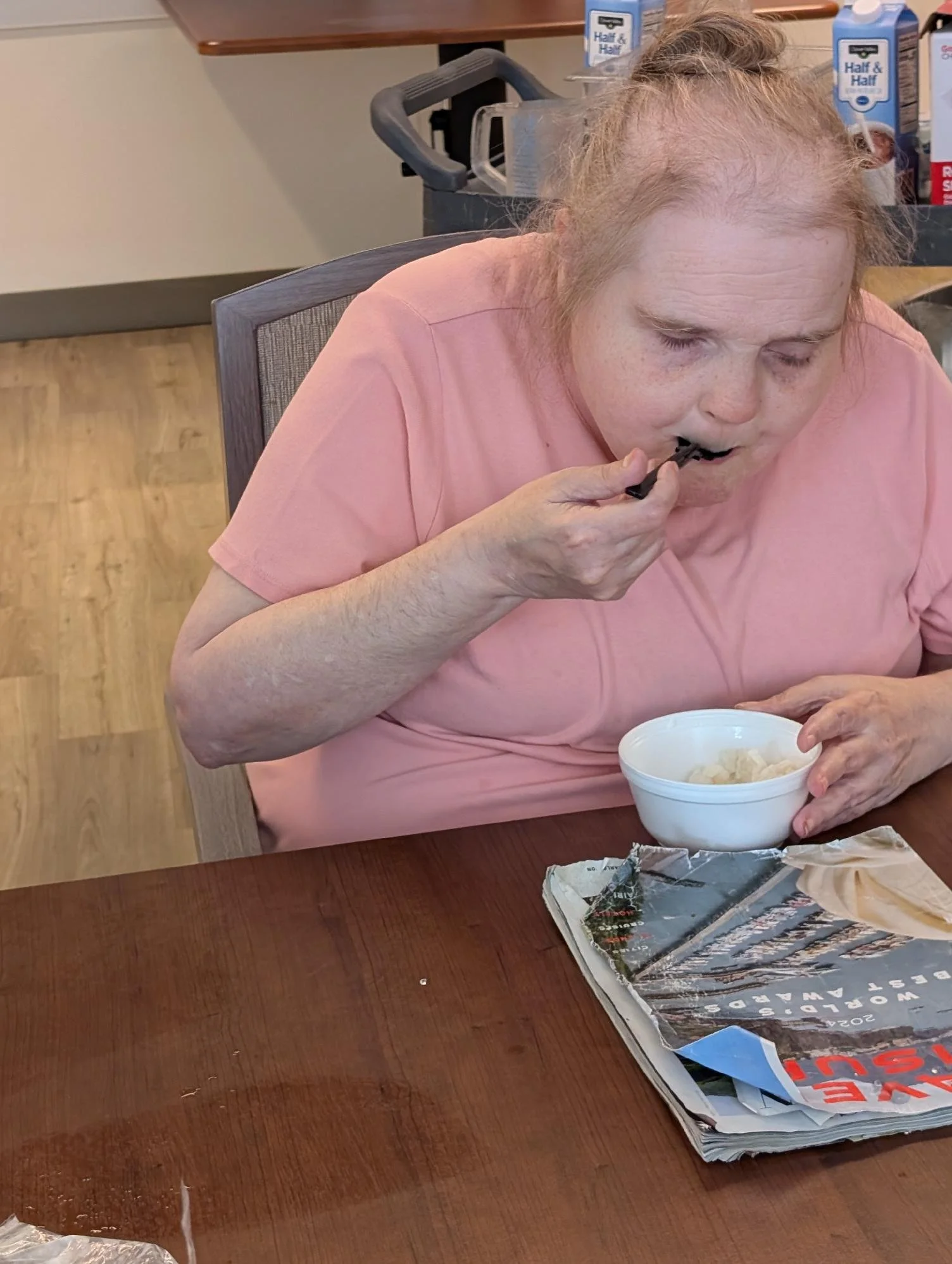 A woman with light skin, red hair, and freckles is sitting at a wooden table eating from a white bowl. She is wearing a pink shirt and using chopsticks. There is a stack of newspapers on the table, and in the background, there are cartons of Half & H