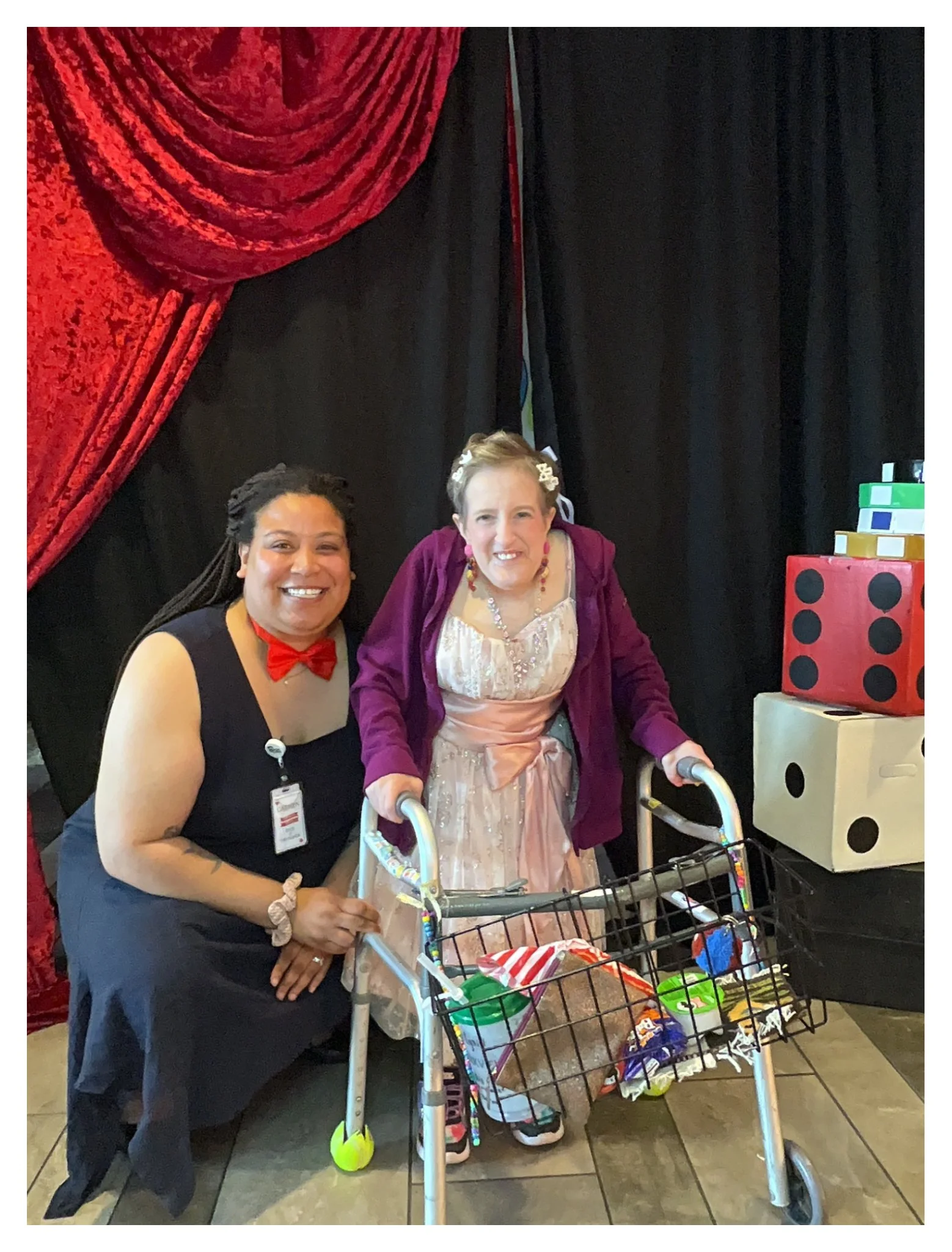 Two women smiling with a woman in a wheelchair in front of a red curtain backdrop, with a display of large colorful dice and a shopping cart filled with snacks in front.