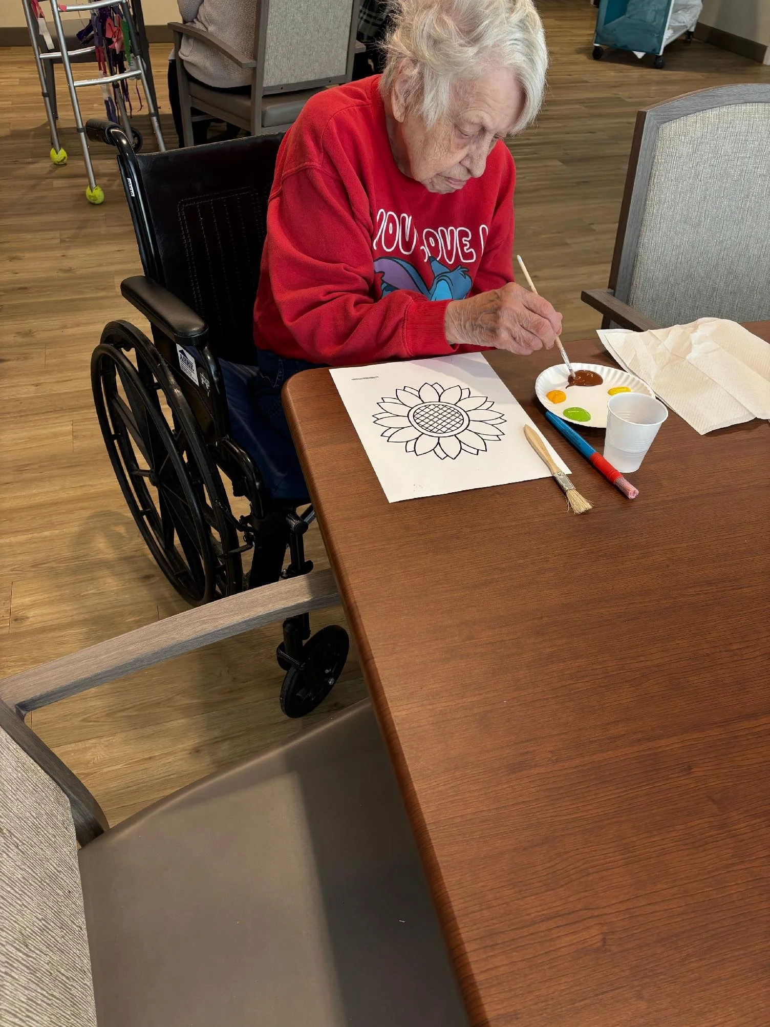 An elderly woman with white hair painting a sunflower outline on paper at a table in a care facility. She is using brown paint and has a paper plate with various colors on it.