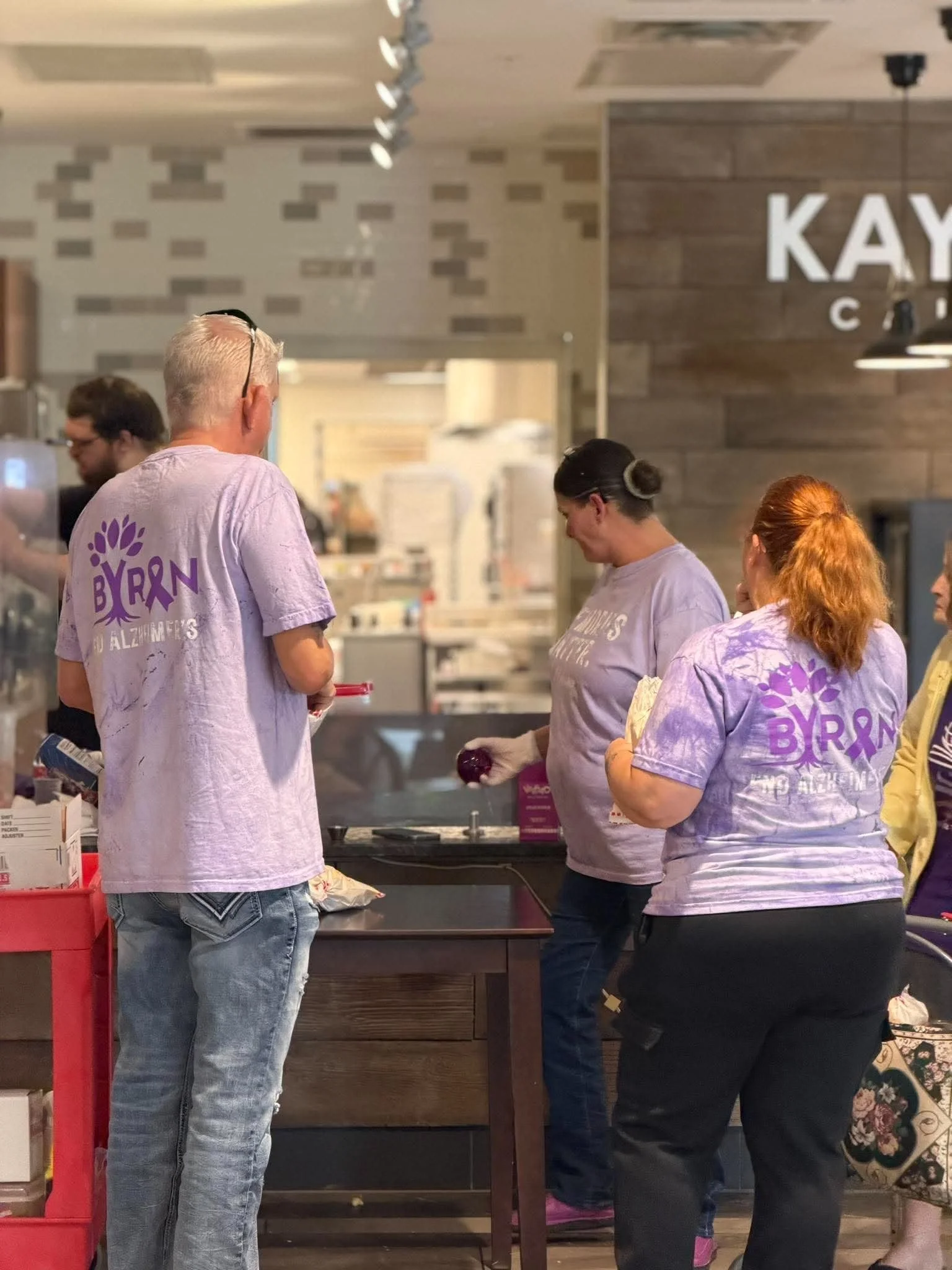 Four people at a counter, with three women and one man. The woman on the right has red hair and is wearing a tie-dye shirt. The woman in the middle has dark hair in a bun. The man has gray hair and is wearing a light pink shirt with purple text. The 