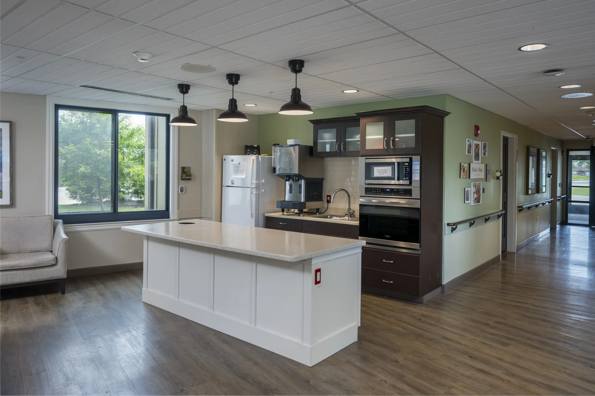An indoor kitchen area with a white island counter, dark brown cabinets, a microwave, oven, and a refrigerator. There are three black pendant lights hanging from the ceiling, and large windows letting in natural light.