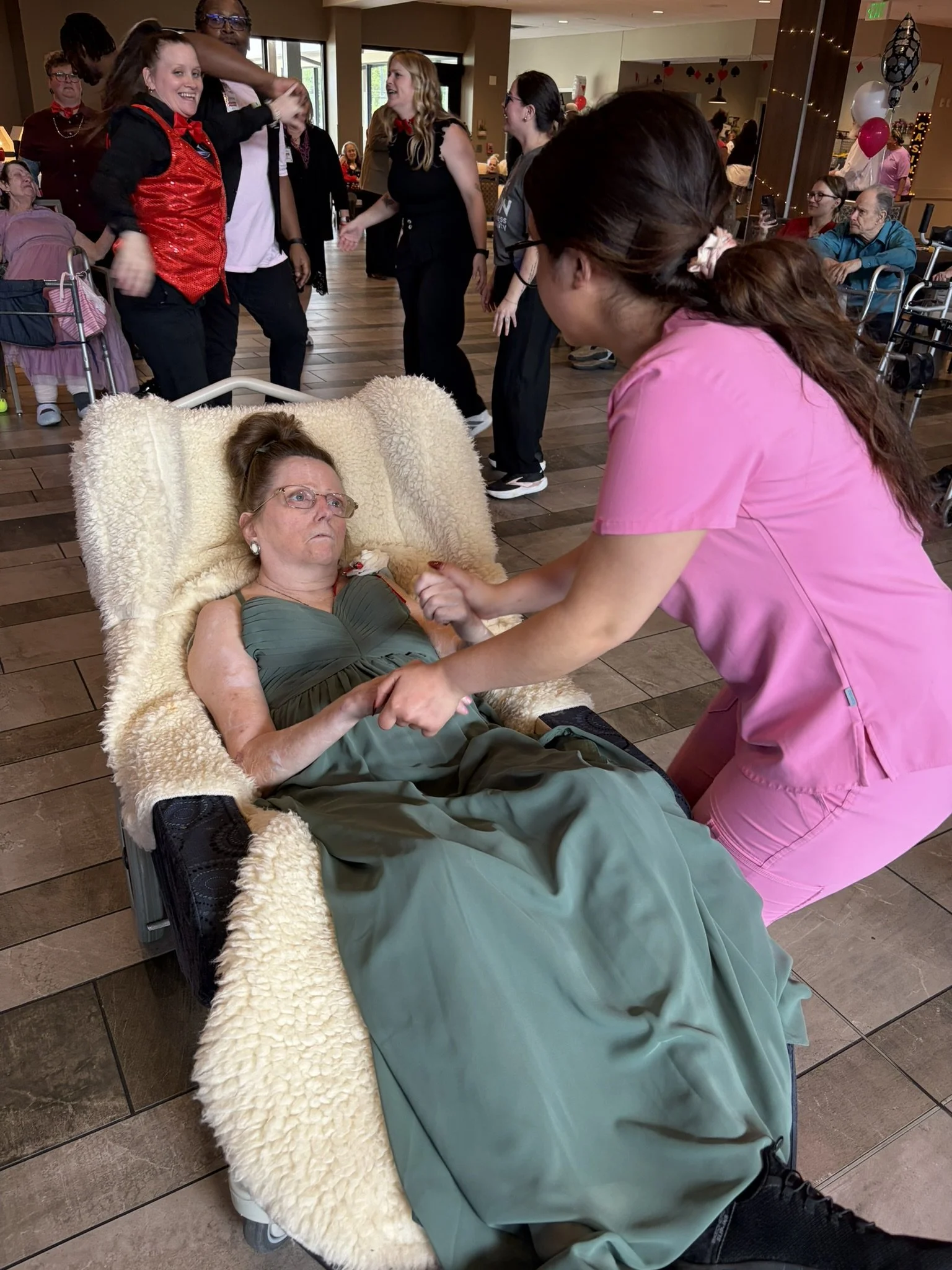 A group of people dancing together in a room decorated for a celebration. In the foreground, a woman with glasses laying on a reclining chair with a fluffy blanket and a woman in pink scrubs holding her hand. In the background, other people are watch
