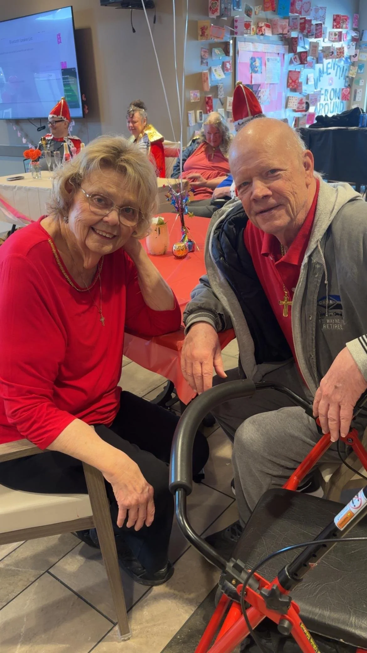 Happy elderly woman and elderly man with a walker sitting at a decorated table during a Valentine's Day celebration with people in festive costumes in the background.