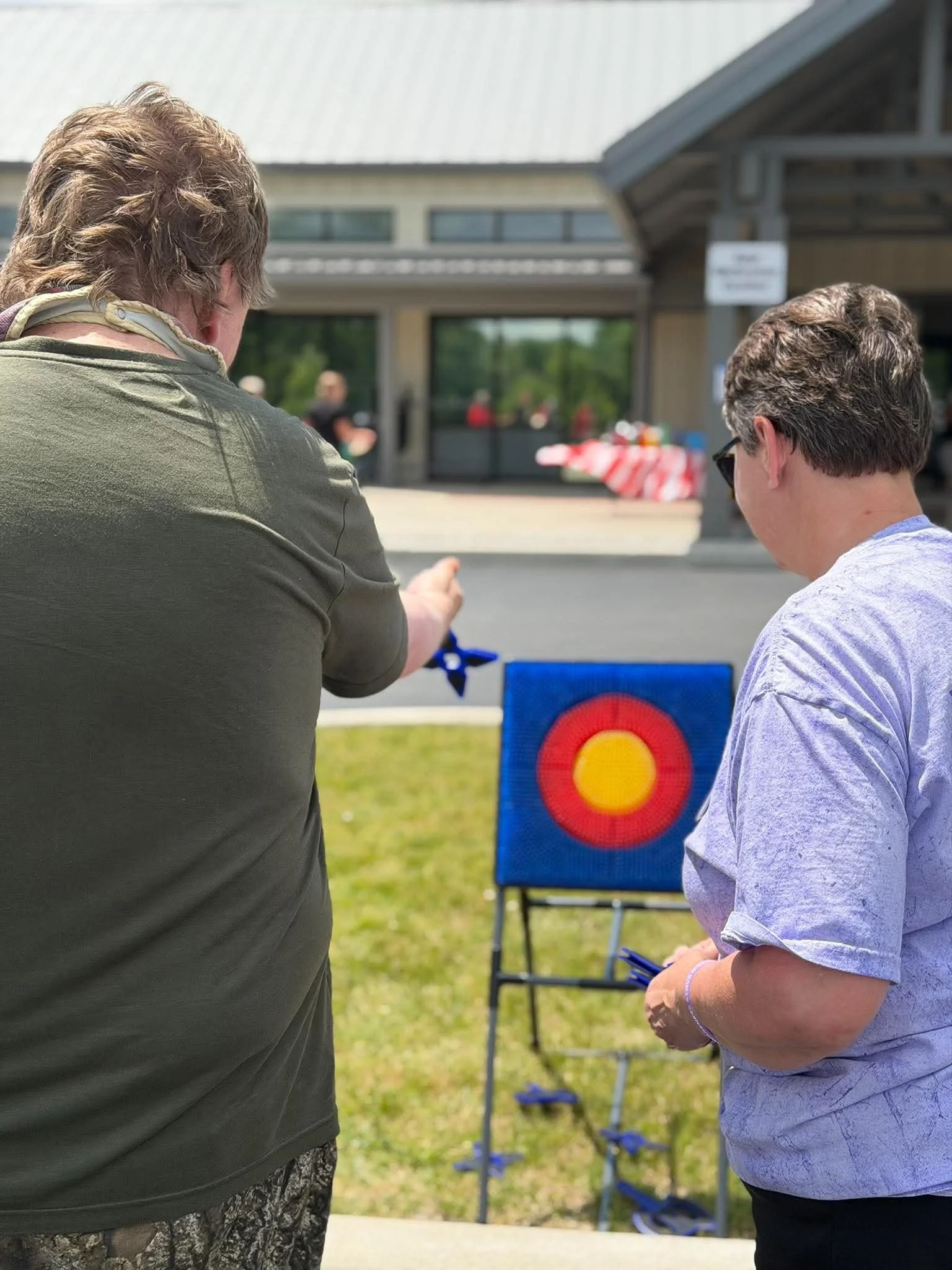 Two people, a woman and a man, stand outdoors near a target used for archery or darts, with trees and a building in the background. The woman wears a green shirt and patterned pants; the man wears a light purple shirt and sunglasses.