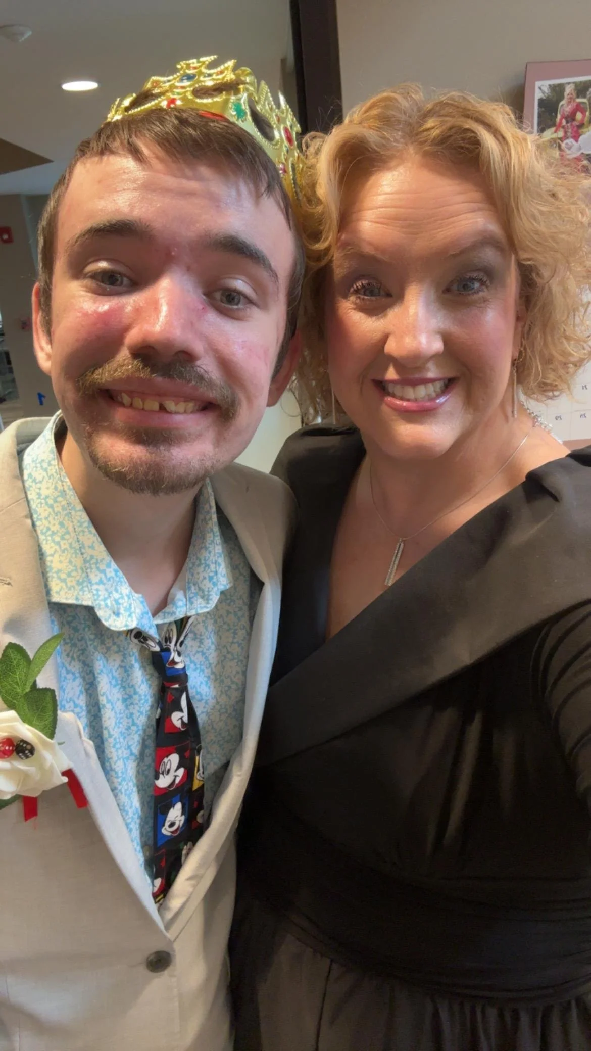 Two people taking a selfie, a young man wearing a gold crown and a woman with curly blonde hair, both smiling at the camera inside a building.
