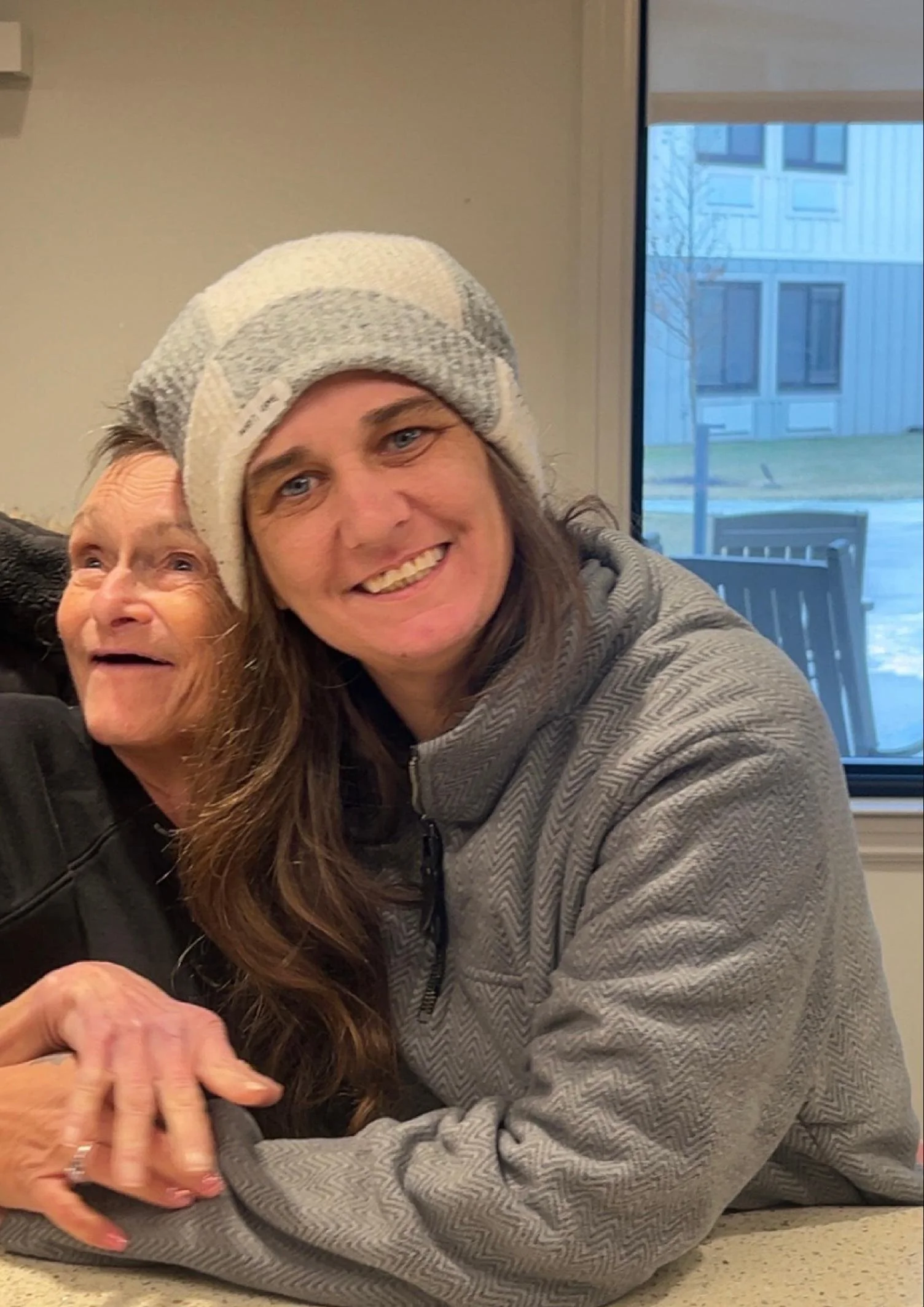 Two women, one older with grey hair and one younger with brown hair, embracing and smiling near a window with a building outside.