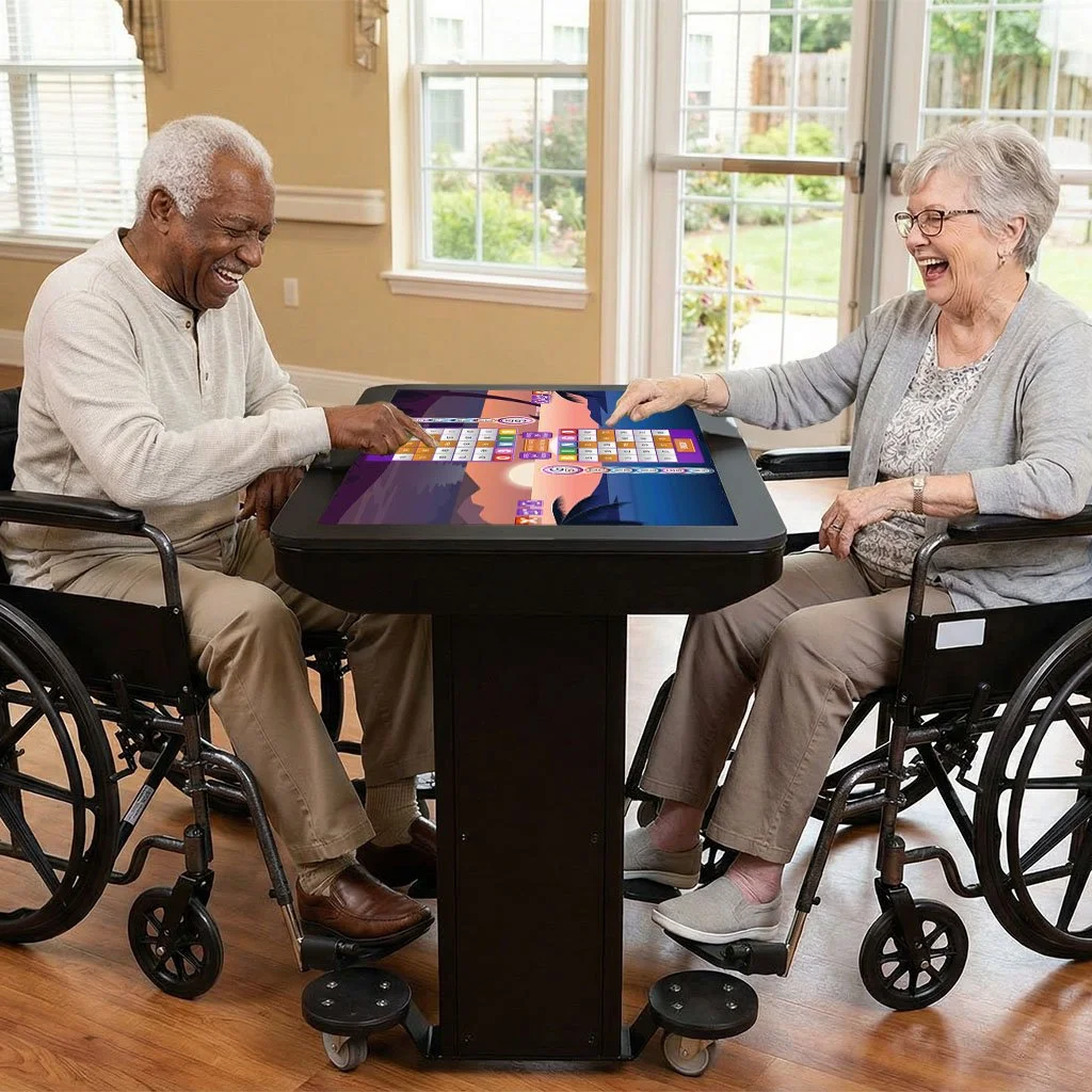 Two elderly people, a man and a woman, sitting in wheelchairs at a table, playing a digital board game on a touchscreen table, smiling and laughing.