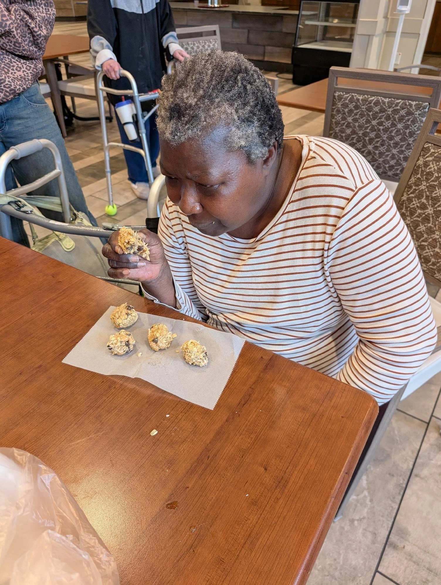 A person with gray hair and a striped shirt sitting at a wooden table, holding a cookie, with more cookies on parchment paper in front of them. Other people and chairs are visible in the background of the restaurant or cafe.