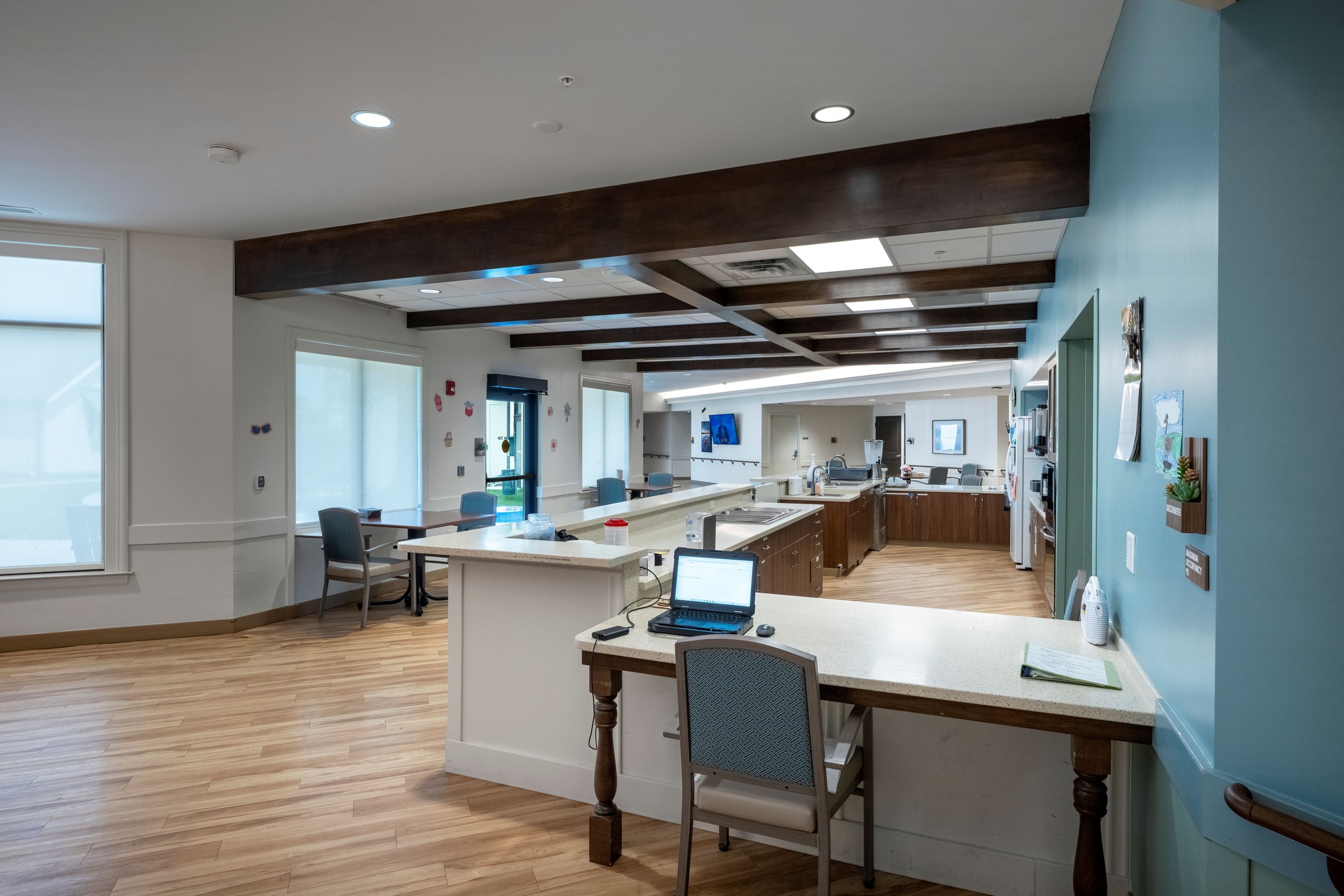 Interior of a communal or dining area with white and teal walls, wood flooring, tables, chairs, and a kitchen with a breakfast bar, sink, and cabinets. Natural light coming through large windows.