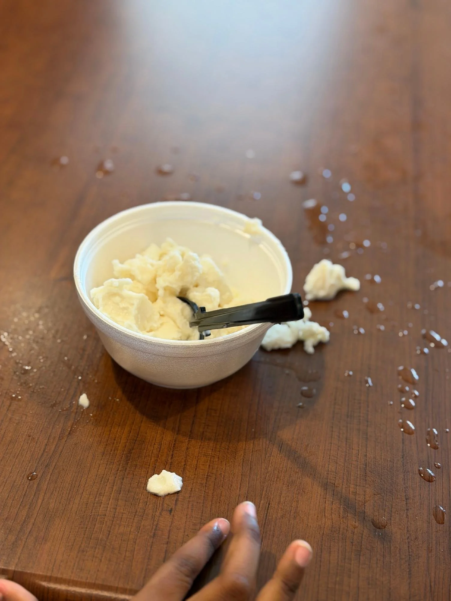 A person's hand is reaching for a piece of ice cream on a wooden table. There is a container of partially eaten vanilla ice cream, with some ice cream and droplets of water scattered around.