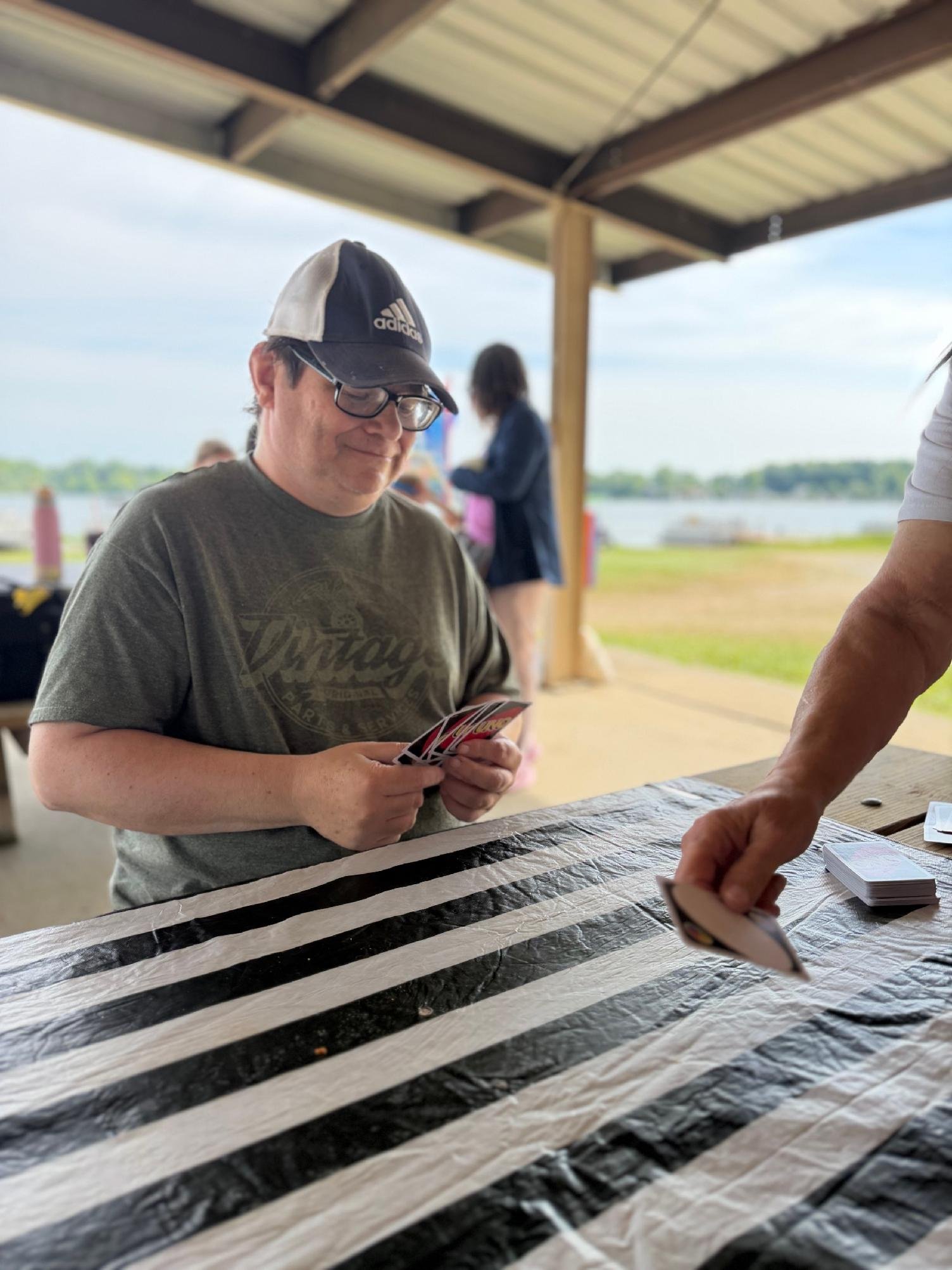 A man wearing glasses and a black and white baseball cap is sitting at a table outdoors, holding playing cards. There are other people in the background near a lake or river, under a covered pavilion with a striped tablecloth on the table.