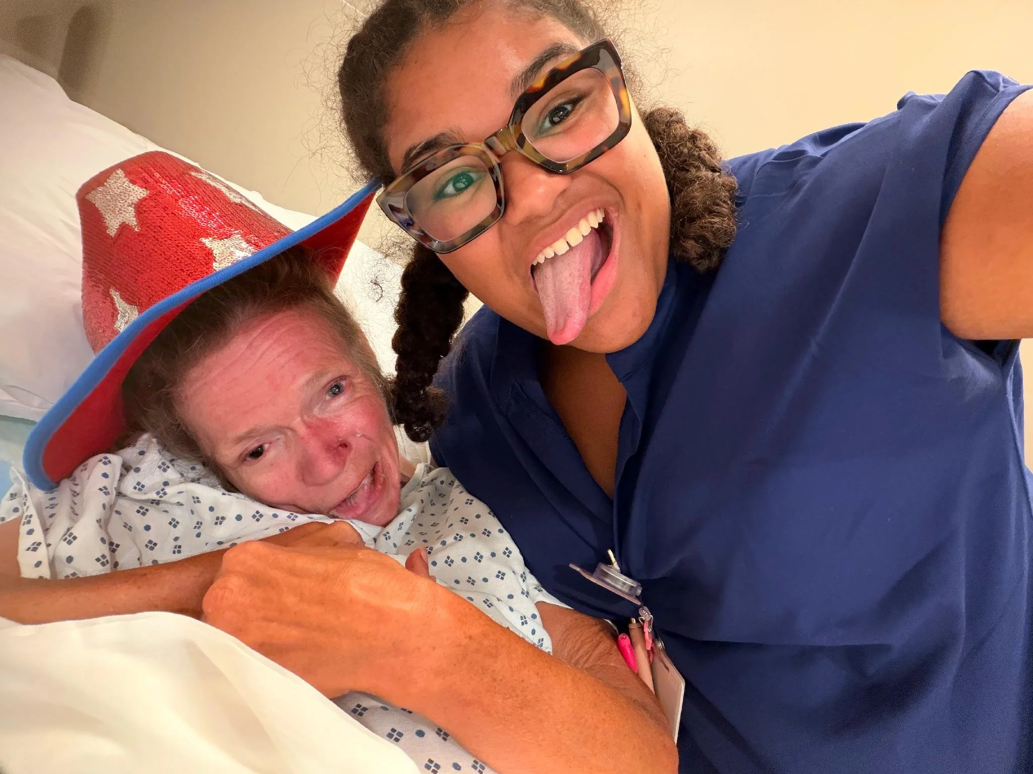 A woman in glasses and a blue shirt is making a playful face with her tongue out, while a person in a hospital gown and a festive hat is lying in a hospital bed. The woman is leaning over the bed, and they appear to be taking a selfie together.