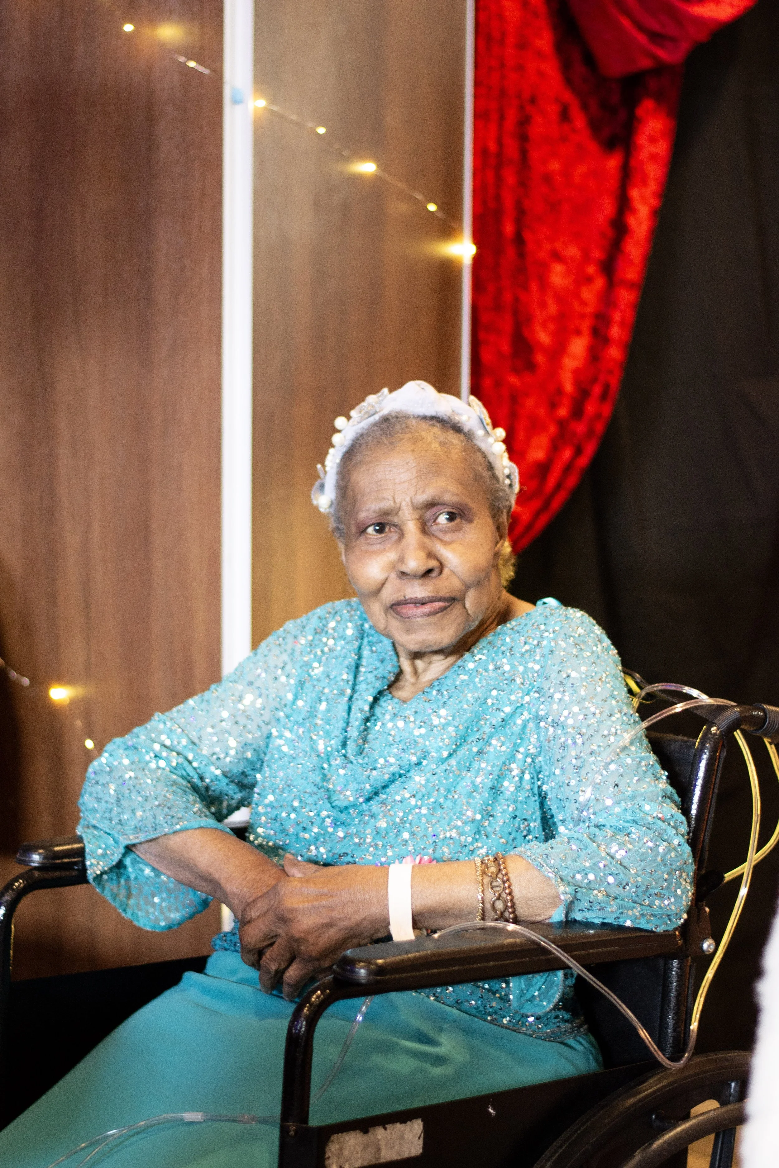 An elderly woman sitting in a wheelchair, wearing a sparkly turquoise dress and pearl headband, with wristbands and a bracelet, in a decorated indoor setting.