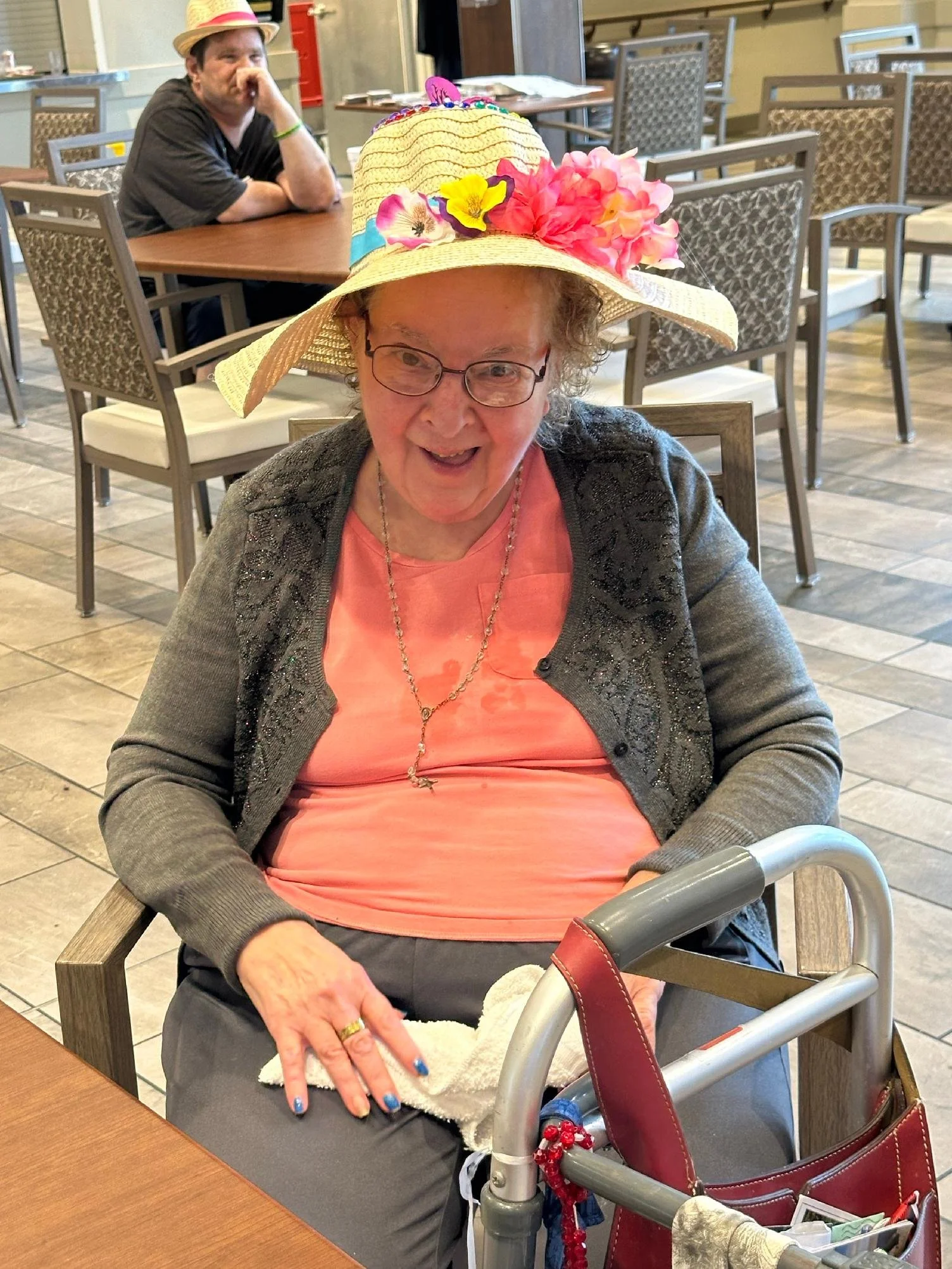 An elderly woman wearing glasses, a pink shirt, a black cardigan with rhinestones, and a large sunhat decorated with colorful flowers is sitting at a table in a restaurant. She appears to be happy and is holding a towel. In the background, a man wear