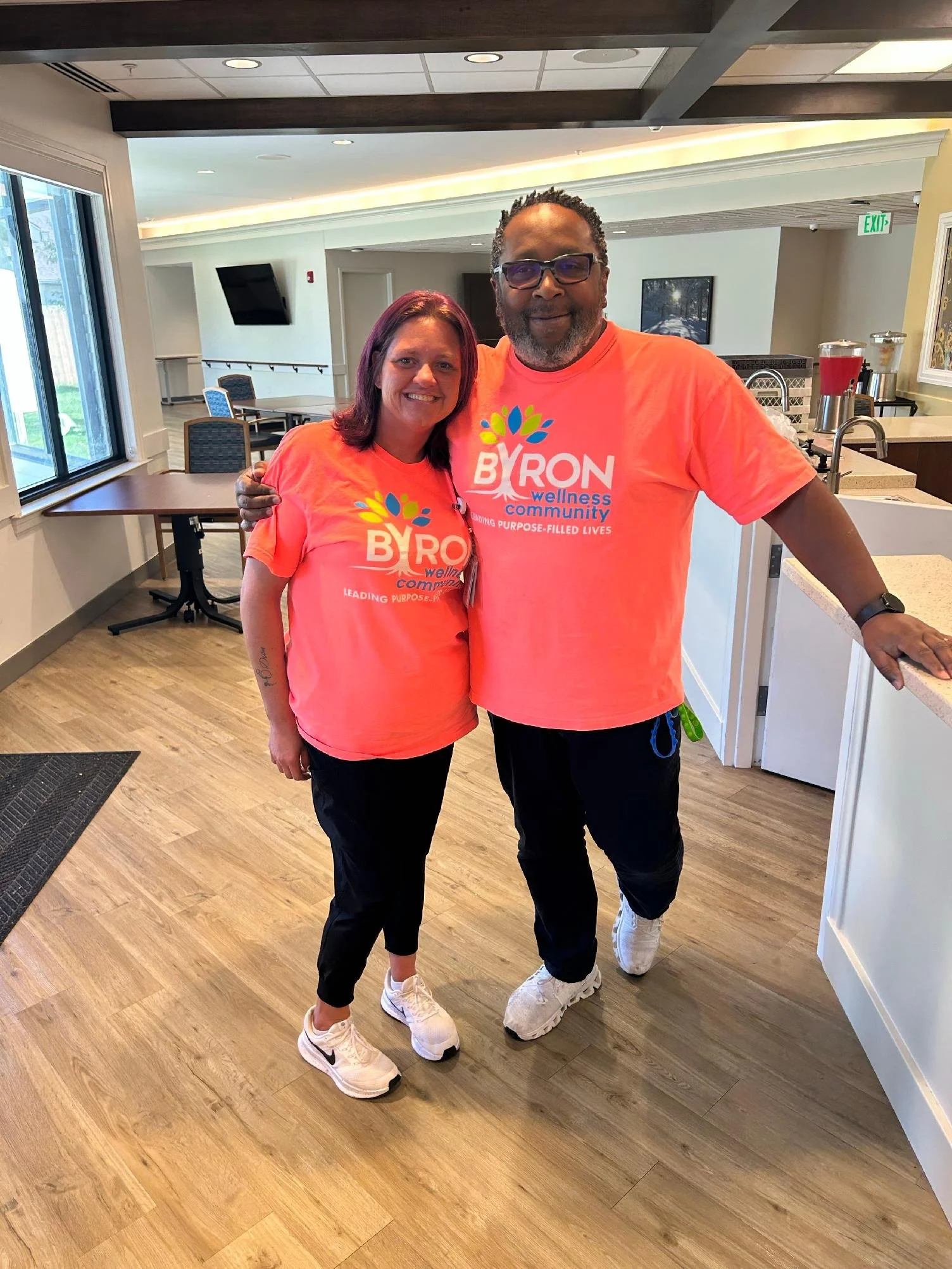 A smiling woman and man wearing matching coral t-shirts with the words 'BYRON wellness community' and a colorful tree logo, standing inside a bright room with wooden floors. They are posing with arms around each other, looking at the camera.