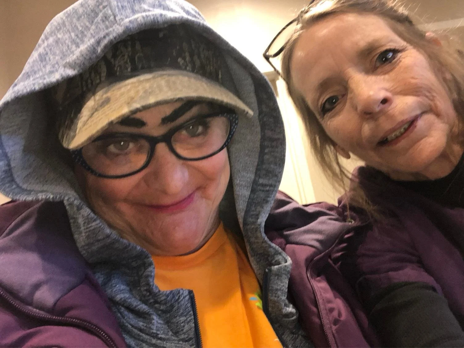 Two women taking a selfie together indoors. The woman on the left is wearing glasses, a hooded sweatshirt, and a baseball cap, with a slight smile. The woman on the right has shoulder-length hair and is smiling. They appear to be close to each other.