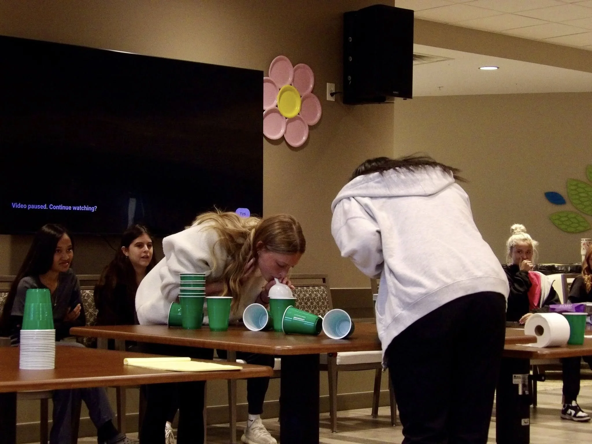 Girls sitting at a table with stacked green and white cups and balls, some cups toppled over, in a room decorated with pink flower-shaped wall decor and green leaf cutouts, possibly during a game or party.