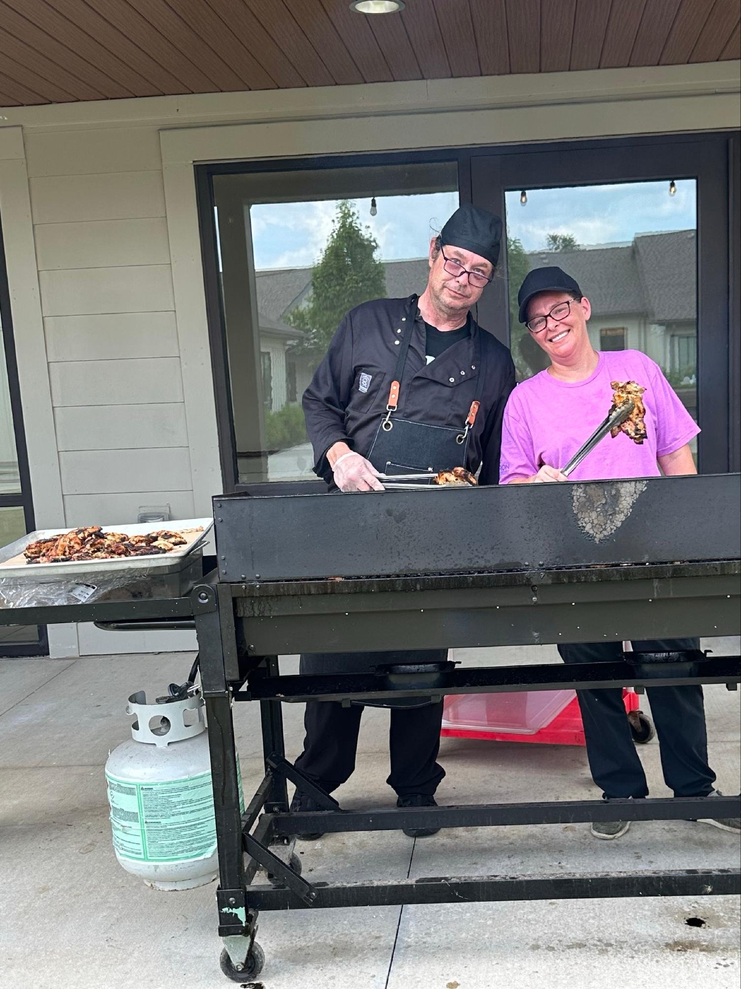 Two people grilling meat outdoors on a barbecue grill, one wearing a black chef hat, glasses, and black apron, and the other wearing a black cap, glasses, and a purple T-shirt, with a house and trees in the background.