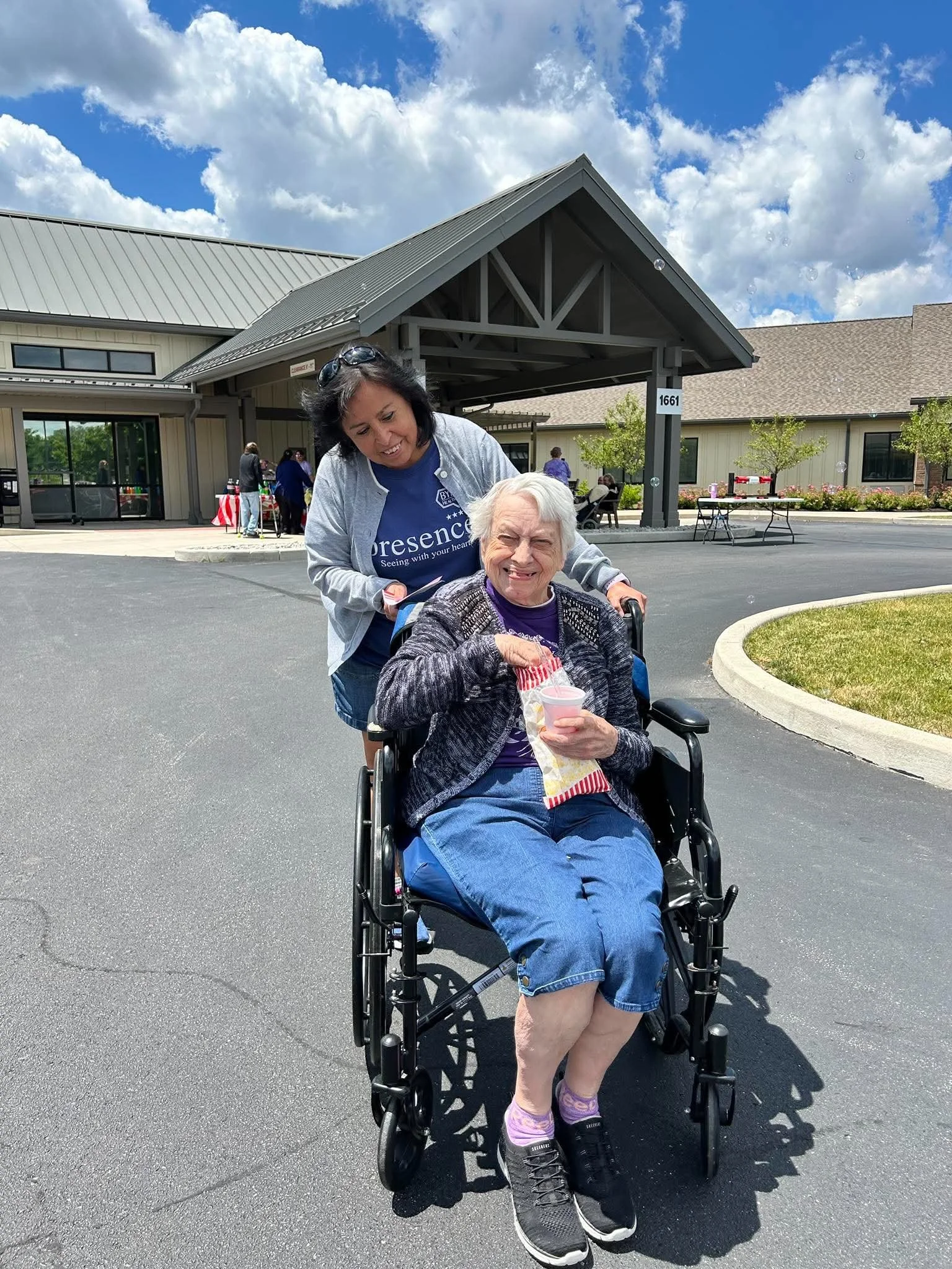 An elderly woman in a wheelchair holding a snack and drink, outdoors in front of a building with a canopy, with a woman standing beside her. The sky is partly cloudy.