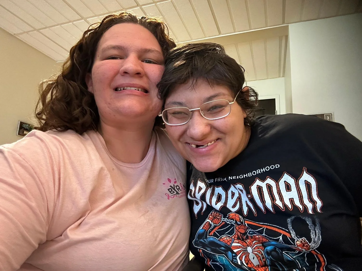 Two people smiling while taking a selfie indoors, one wearing a pink shirt and the other wearing a black T-shirt with a Spider-Man graphic.