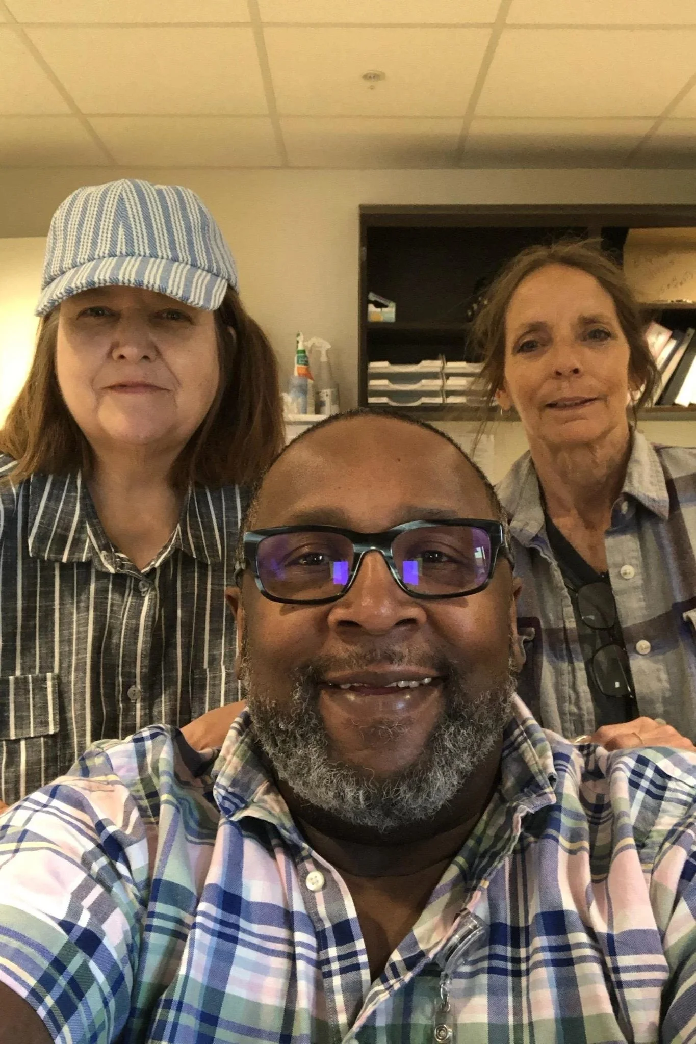 Three people taking a selfie in an indoor setting, with a man in the foreground wearing glasses and a plaid shirt, a woman in the background on the left wearing a striped hat, and another woman to the right wearing a plaid shirt and holding sunglasses, with a bookshelf and cleaning supplies visible in the background.