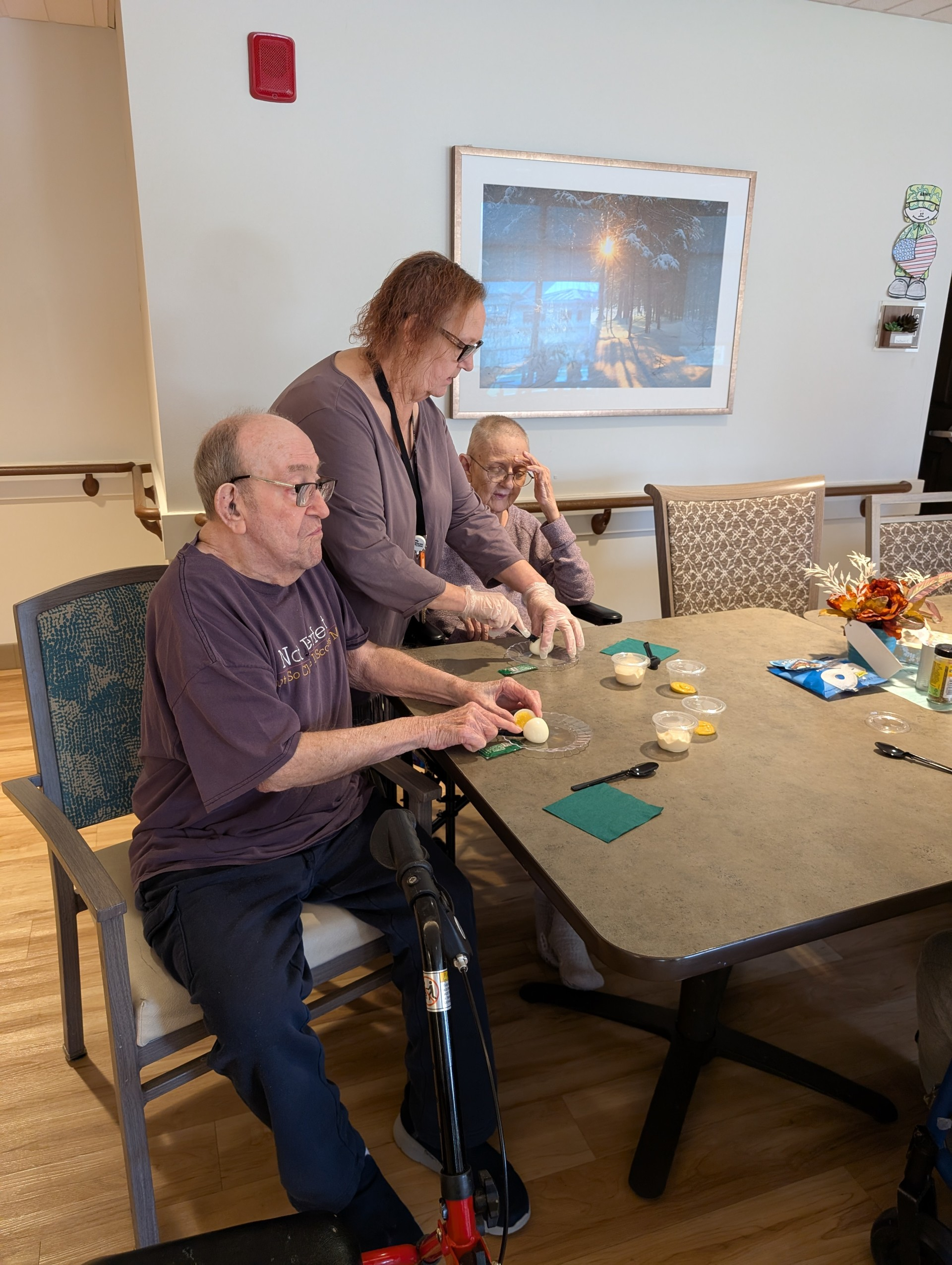 Three elderly people are sitting at a table with various items on it, including containers, papers, and utensils, with a woman standing and assisting them. One person is in a wheelchair. The setting appears to be a social or care facility, with a large framed photograph of a snowy forest on the wall.