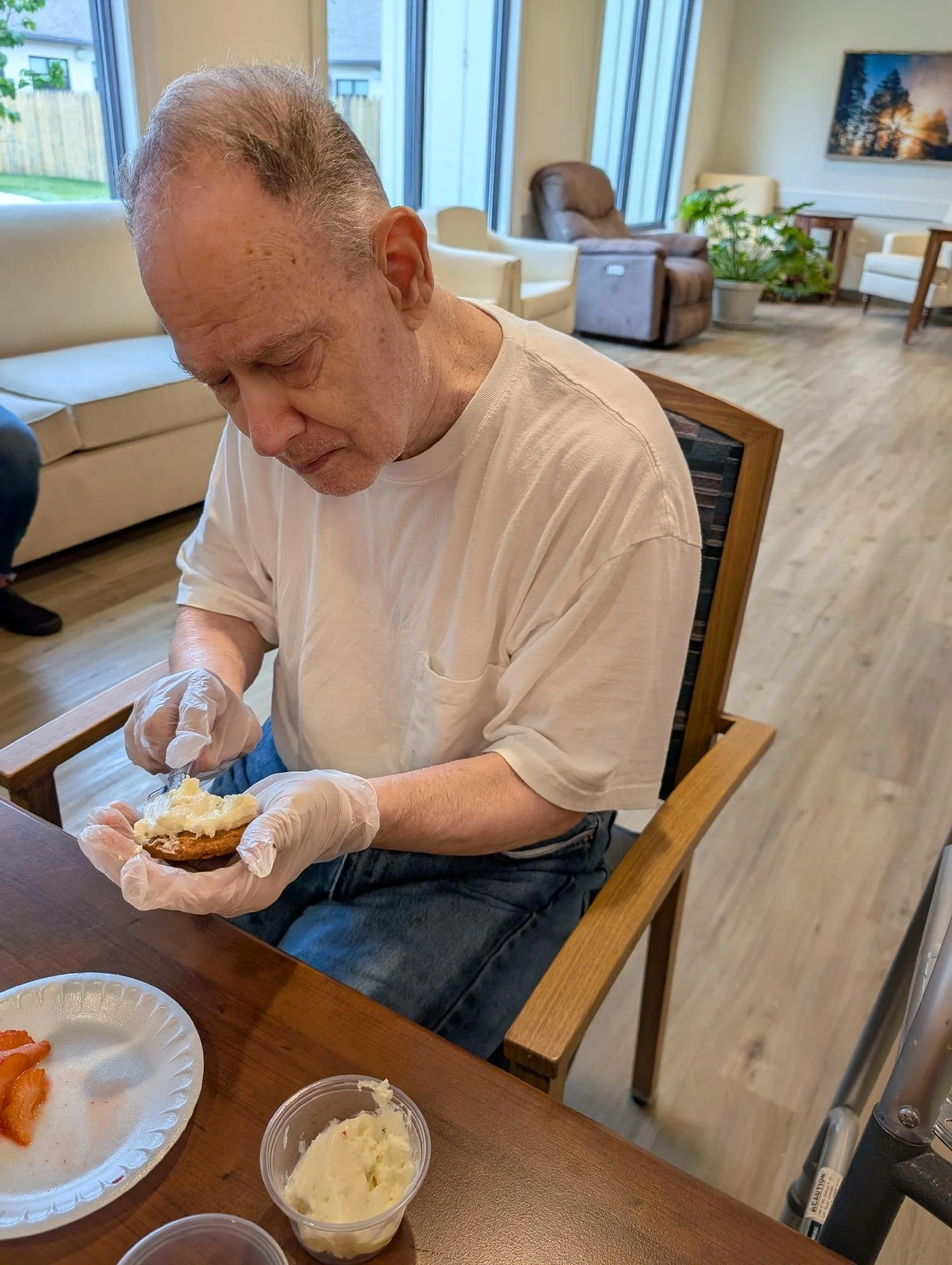 An elderly man with gray hair and wearing a white T-shirt is sitting at a wooden table, spreading butter on bread. The table has various food items like mashed potatoes and sliced carrots. In the background, there are large windows, comfortable chair