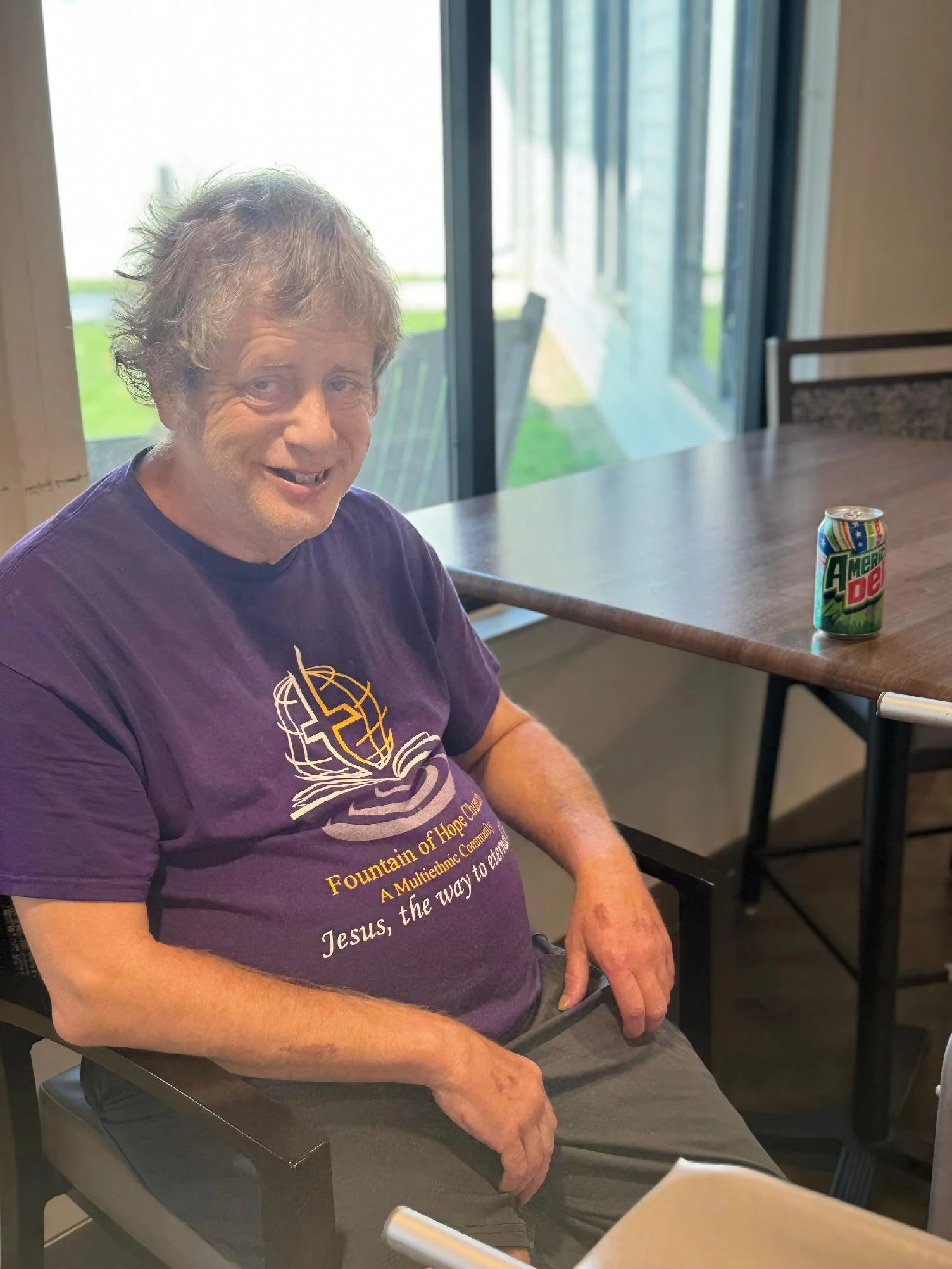 A smiling middle-aged man with gray hair sitting at a table indoors, wearing a purple T-shirt supporting a religious charity event, with a can of Mountain Dew soda on the table beside him.