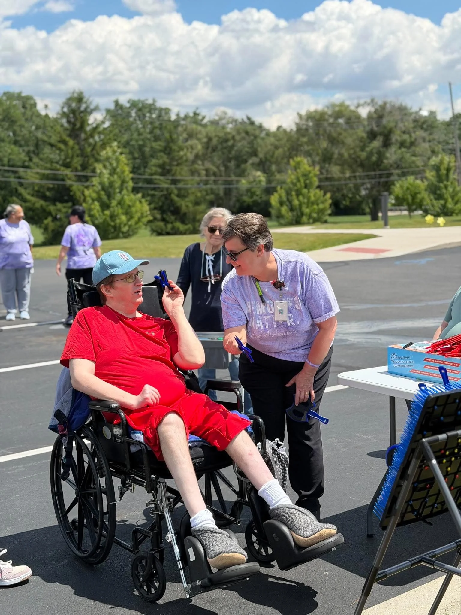 A man in a wheelchair wearing a red shirt and a blue cap is talking to a woman in a purple shirt. There are several women standing in the background on a parking lot, with trees and clouds in the sky.