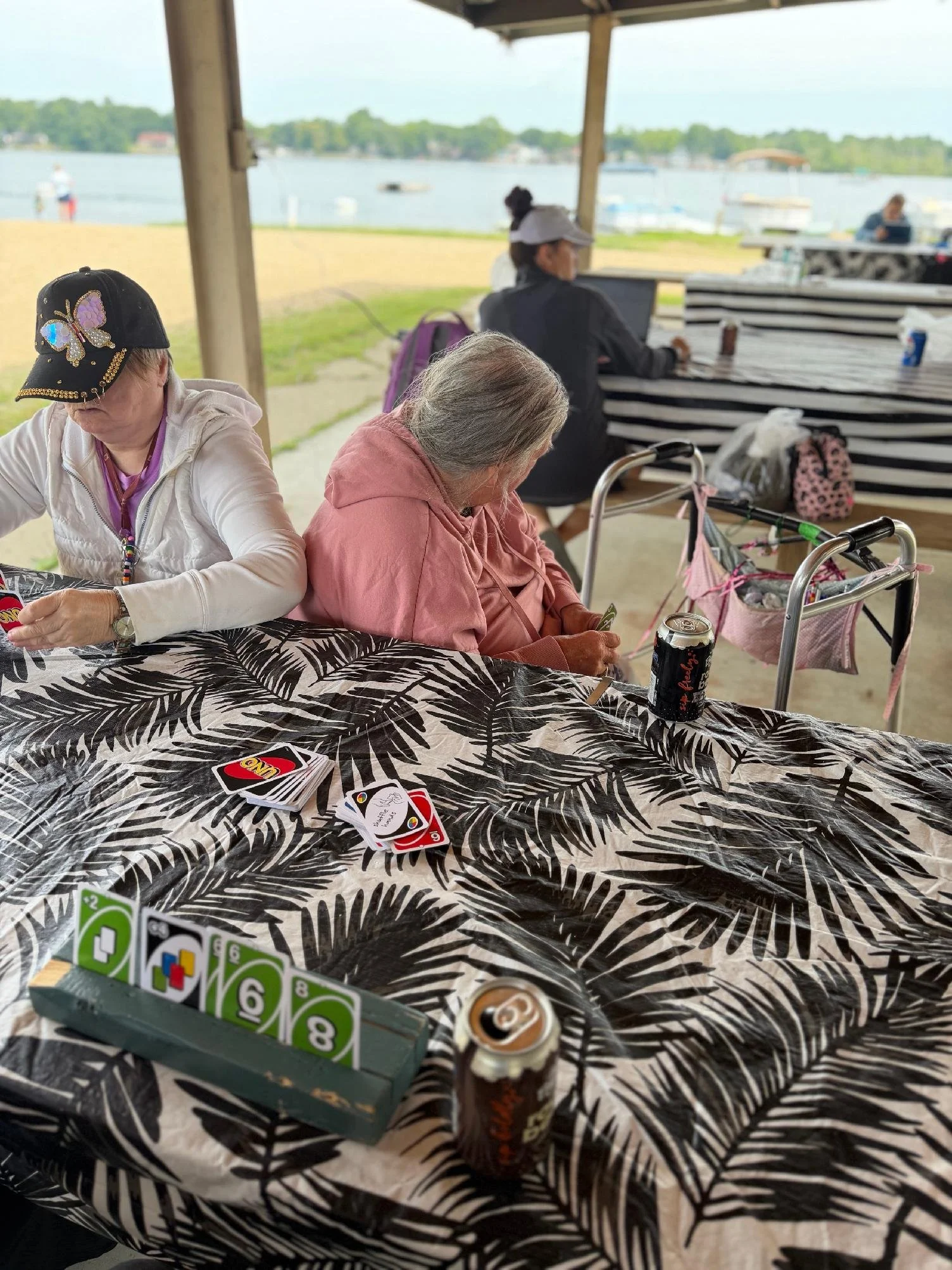 People playing a card game at a table near a lakeside with boats and trees in the background.