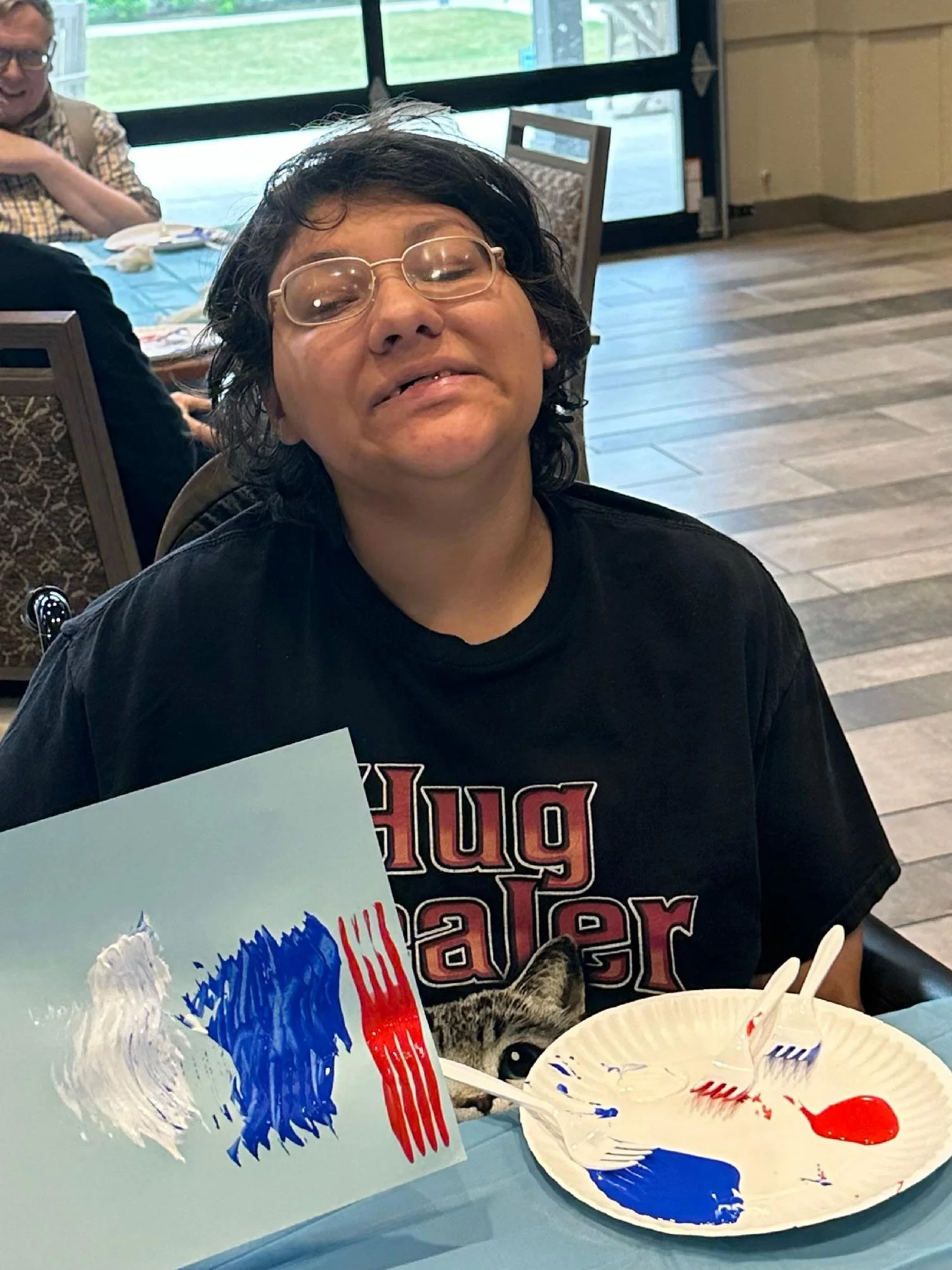 Person with glasses and wavy black hair sitting at a table in a restaurant, with a painted plate and a sign with red, white, and blue paint in front, and blurry background of other people and windows.