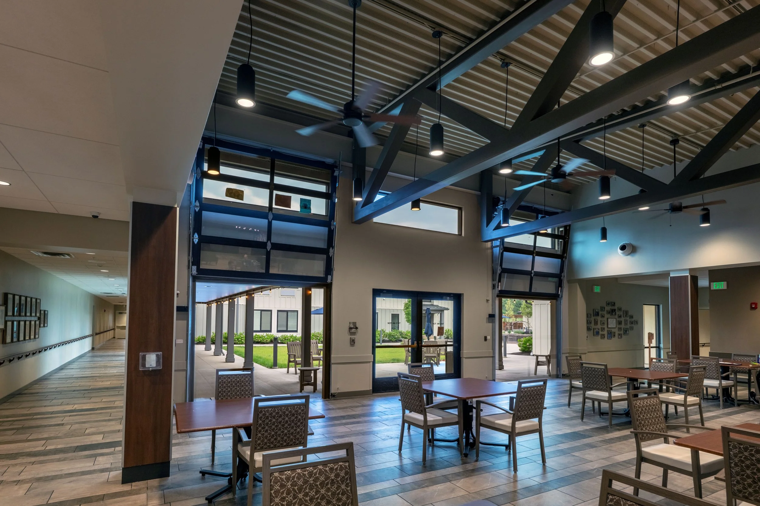 Interior of a modern building with a high ceiling, ceiling fans, hanging lights, large windows, and seating areas with tables and chairs. Outside view shows a patio with benches and greenery.