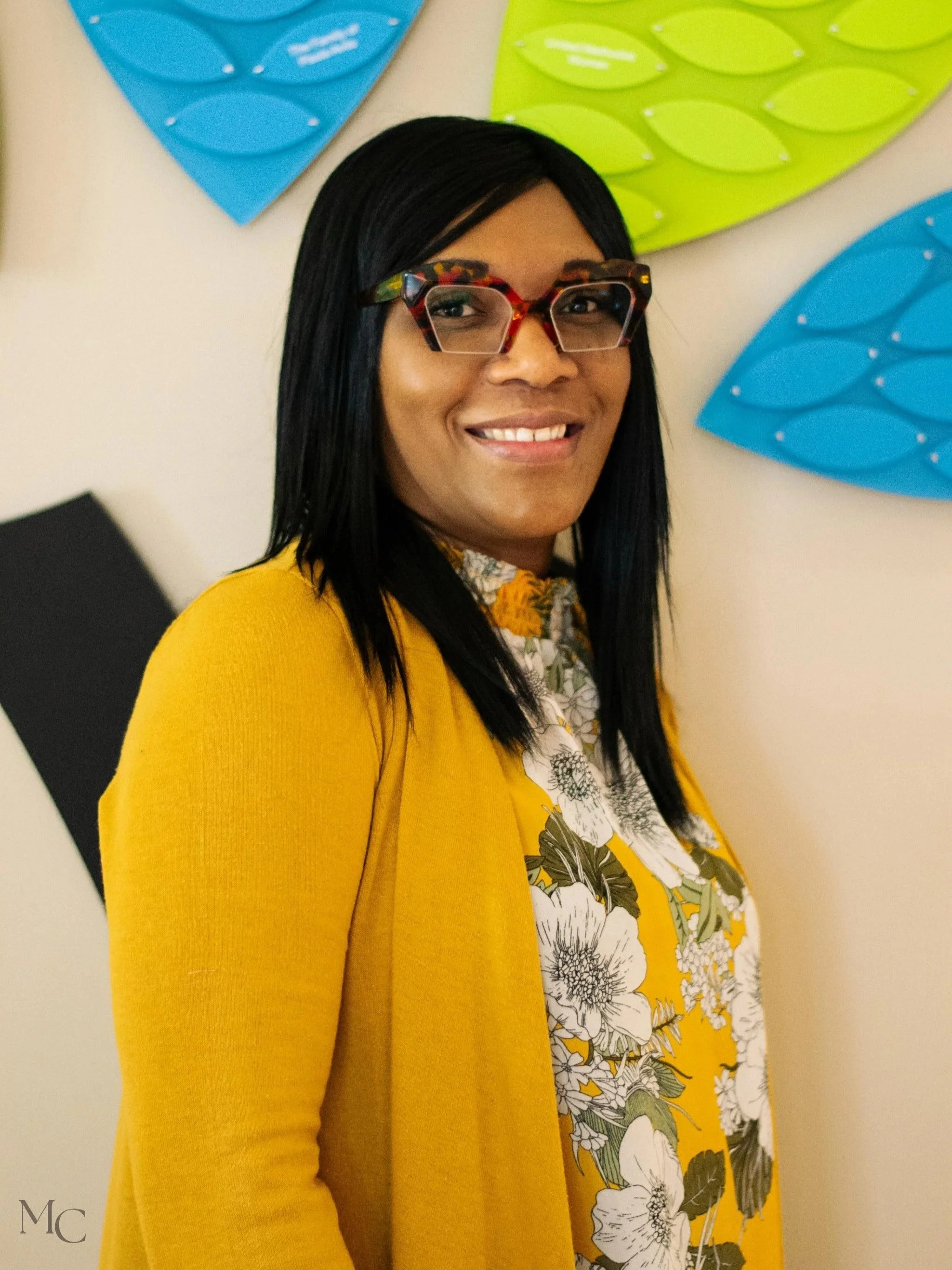 Portrait of a woman with long black hair, wearing colorful glasses, a yellow cardigan, and a floral blouse, smiling in front of decorative blue and green wall hangings.