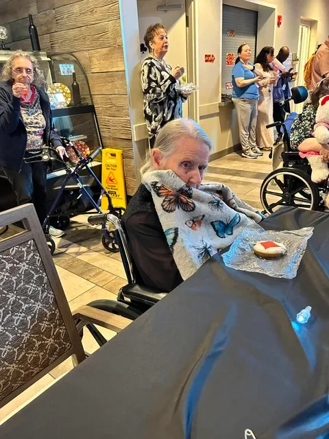 Elderly woman sitting in a wheelchair with a butterfly-patterned blanket, holding a cookie. Others are standing and sitting in the background, some with wheelchairs, in a room with wooden walls and a dark table in the foreground.