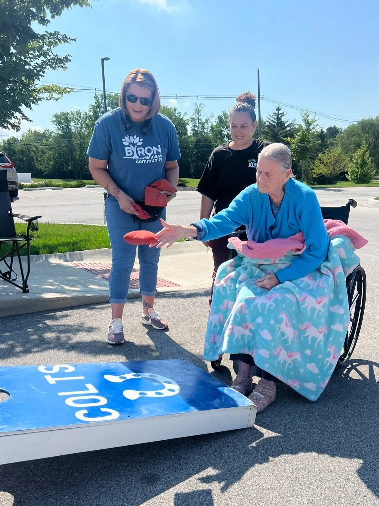 Three women engage in a game of cornhole outdoors on a sunny day. One woman in a wheelchair is throwing a beanbag, while two women stand nearby smiling. They are on a paved area with a grassy background and trees.