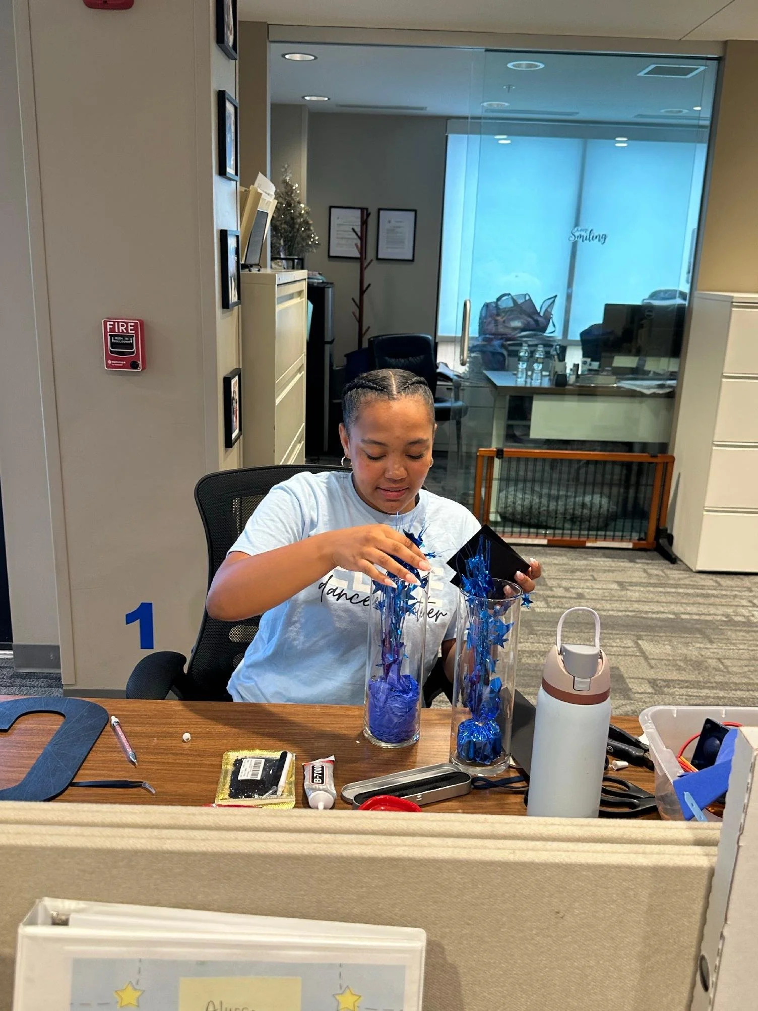 A young girl with braided hair sitting at a desk, decorating a tall glass jar with blue tinsel. The desk has various craft supplies and a white water bottle. There is a partition in front of her and a glass wall behind her with a view of an office wi