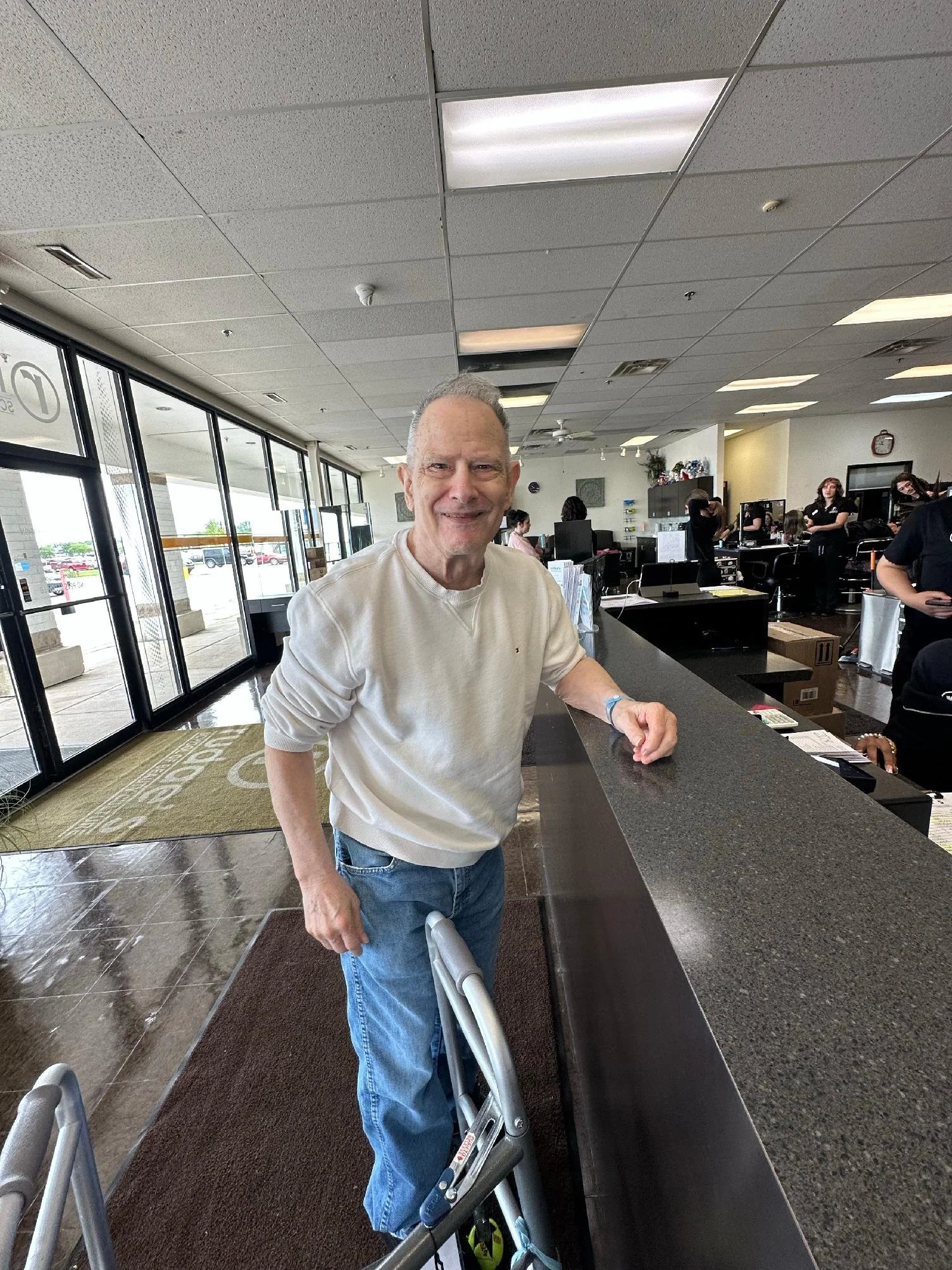 A smiling older man stands at a check-in counter with a rollator walker at his side, inside a busy office or reception area with several people working behind the counter.